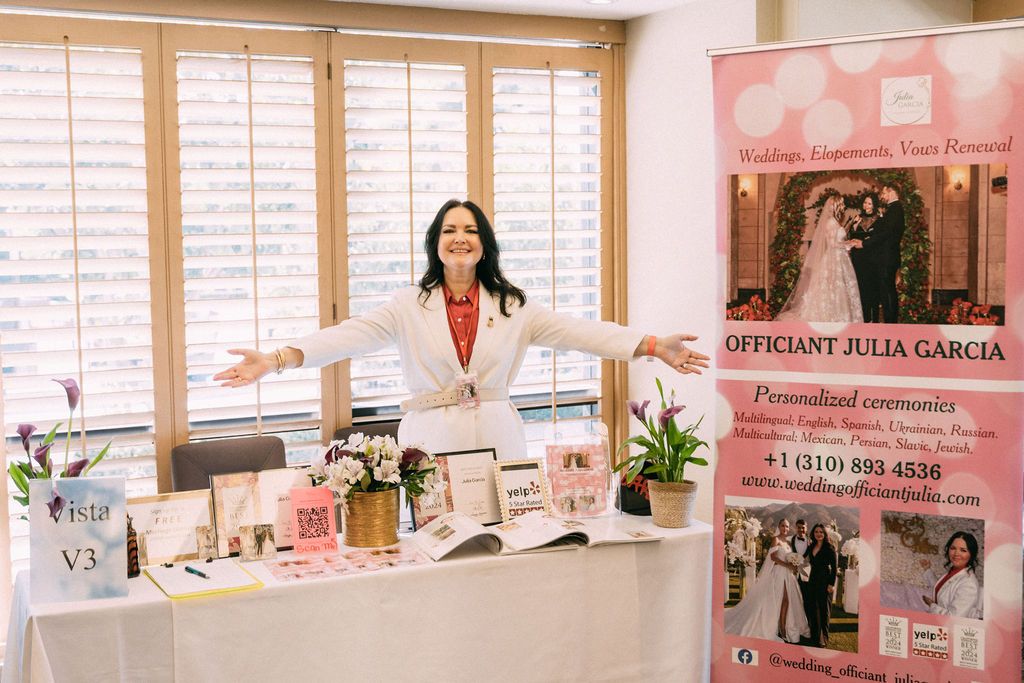 Wedding officiant Julia Garcia posing with open arms at her decorated vendor booth at a bridal expo.