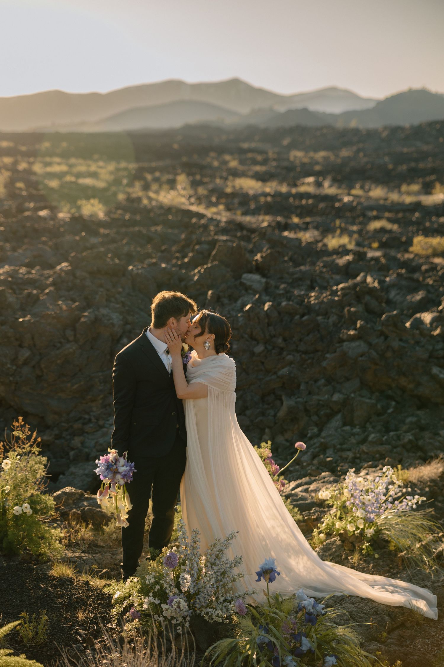 Bride and groom share a romantic kiss at golden hour among volcanic rock landscape with mountain views.