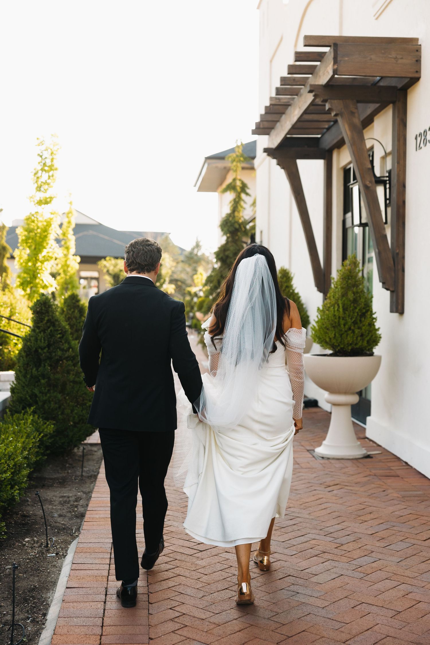 Bride in white gown and veil walking with groom along brick pathway outdoors.