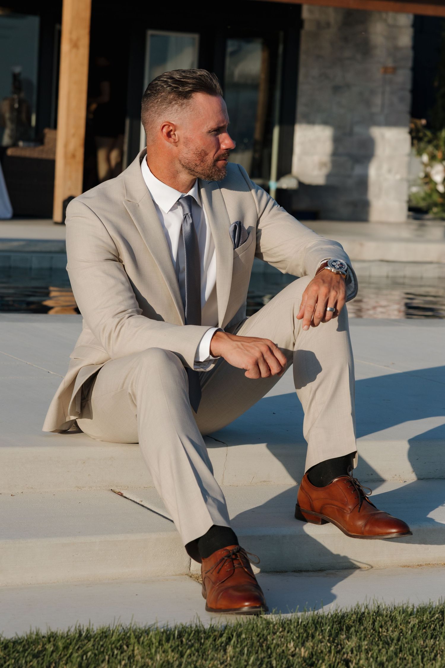 Groom in beige suit and brown dress shoes sitting relaxed poolside at outdoor wedding venue.