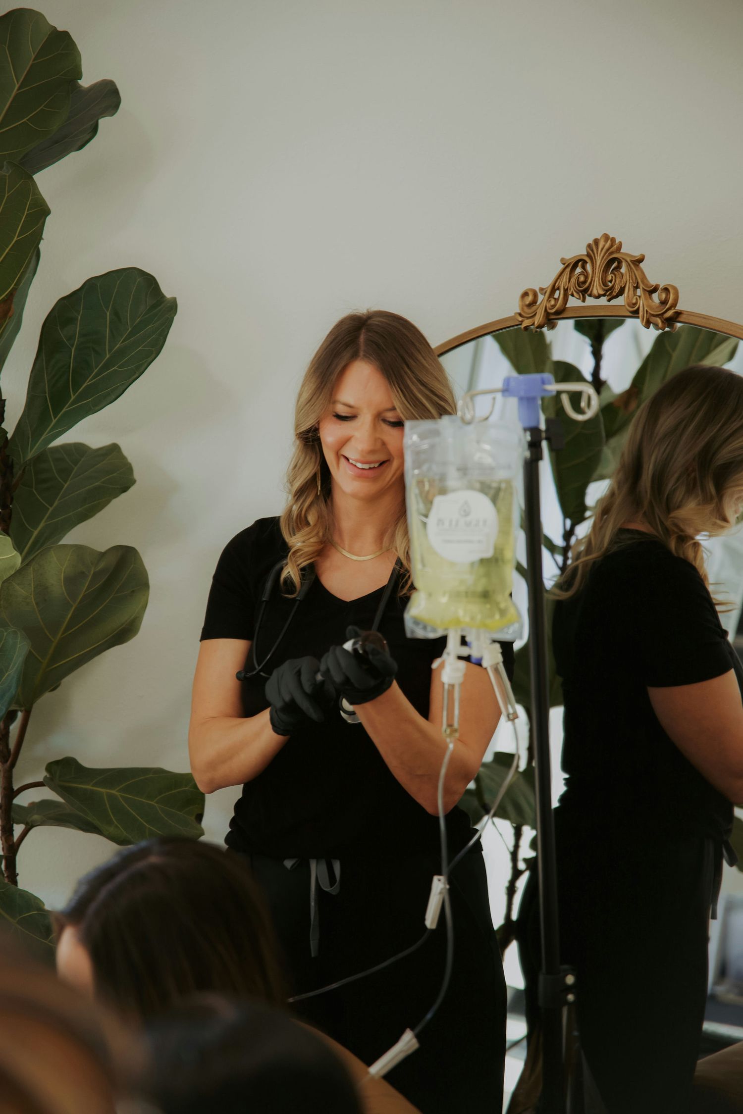 A smiling nurse in black gloves prepares an IV drip bag in a modern wellness clinic with a decorative mirror.