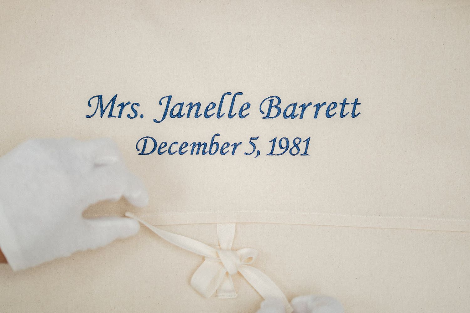 Cream fabric with blue cursive text reading Mrs. Janelle Barrett, December 5, 1981, with a white ribbon bow.