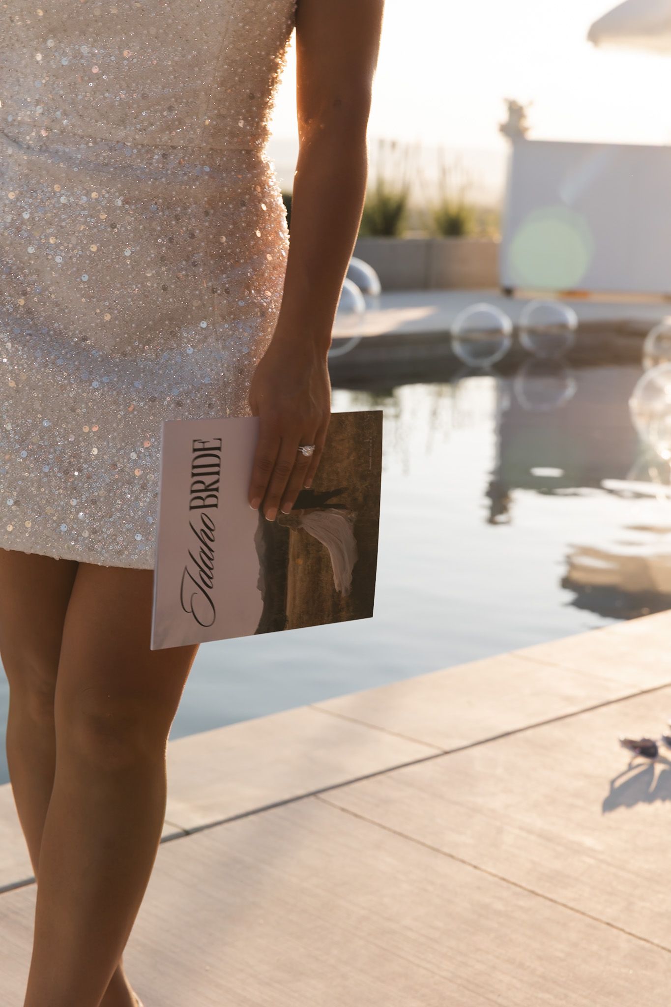 Future bride in sequin mini dress holding card by poolside at sunset.