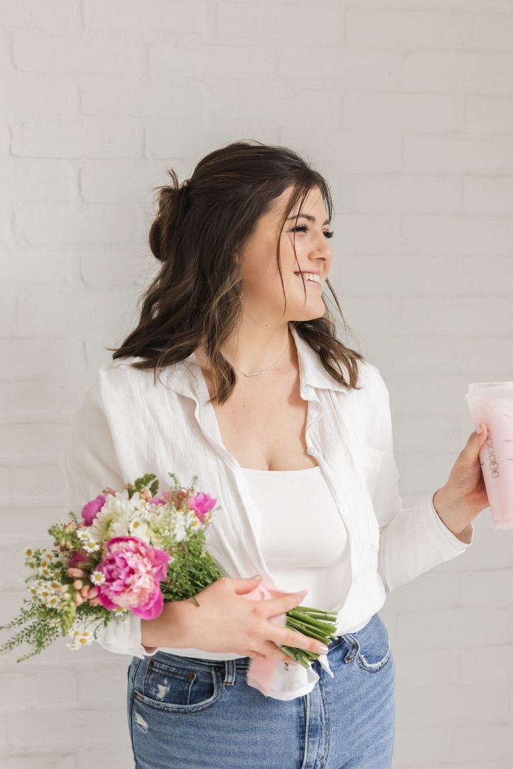 Happy young woman in white blouse holding colorful peony bouquet and pink drink against white brick wall.
