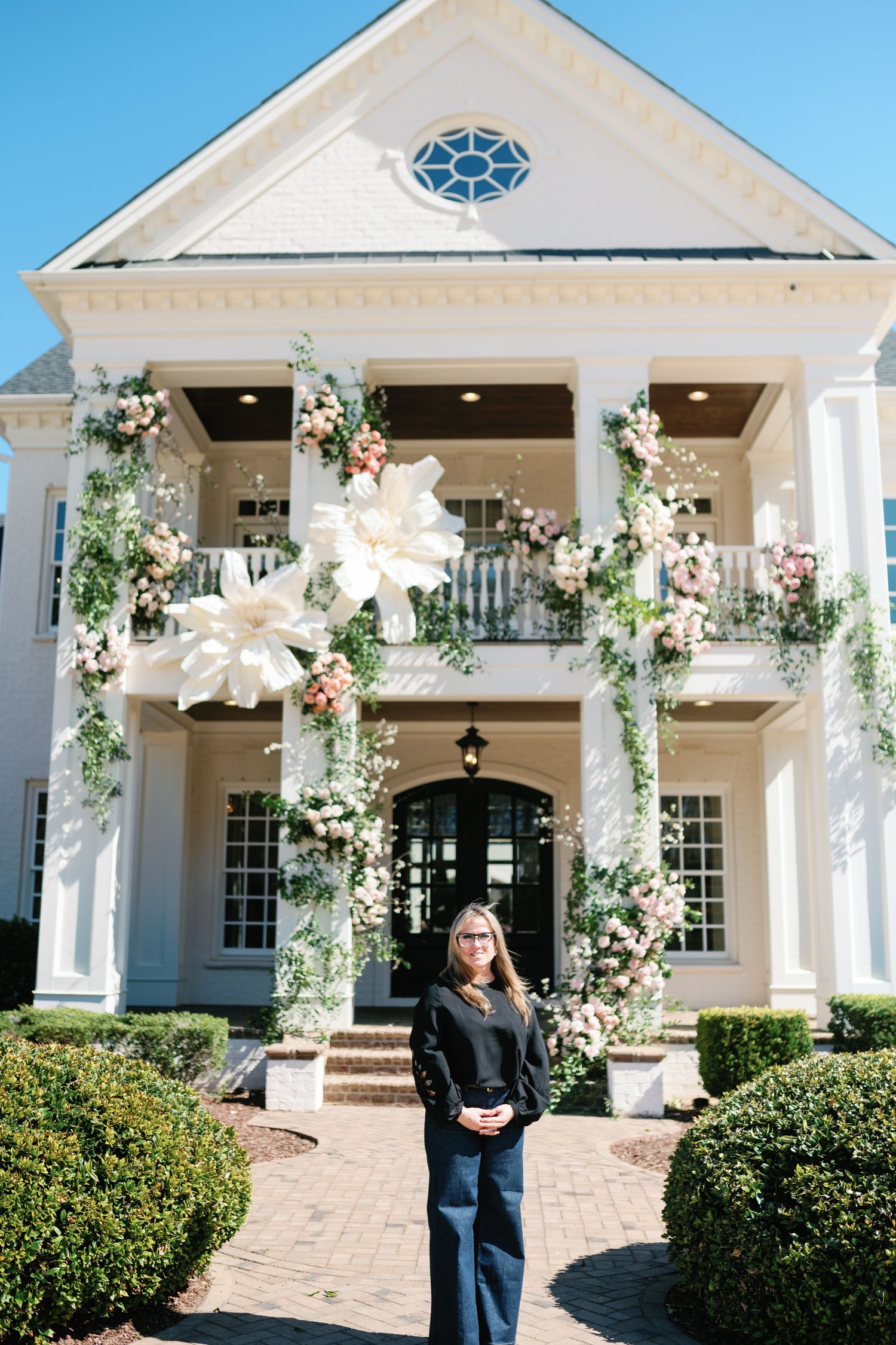 Woman stands before an elegant white colonial wedding venue adorned with lush floral arrangements on columns and balcony.