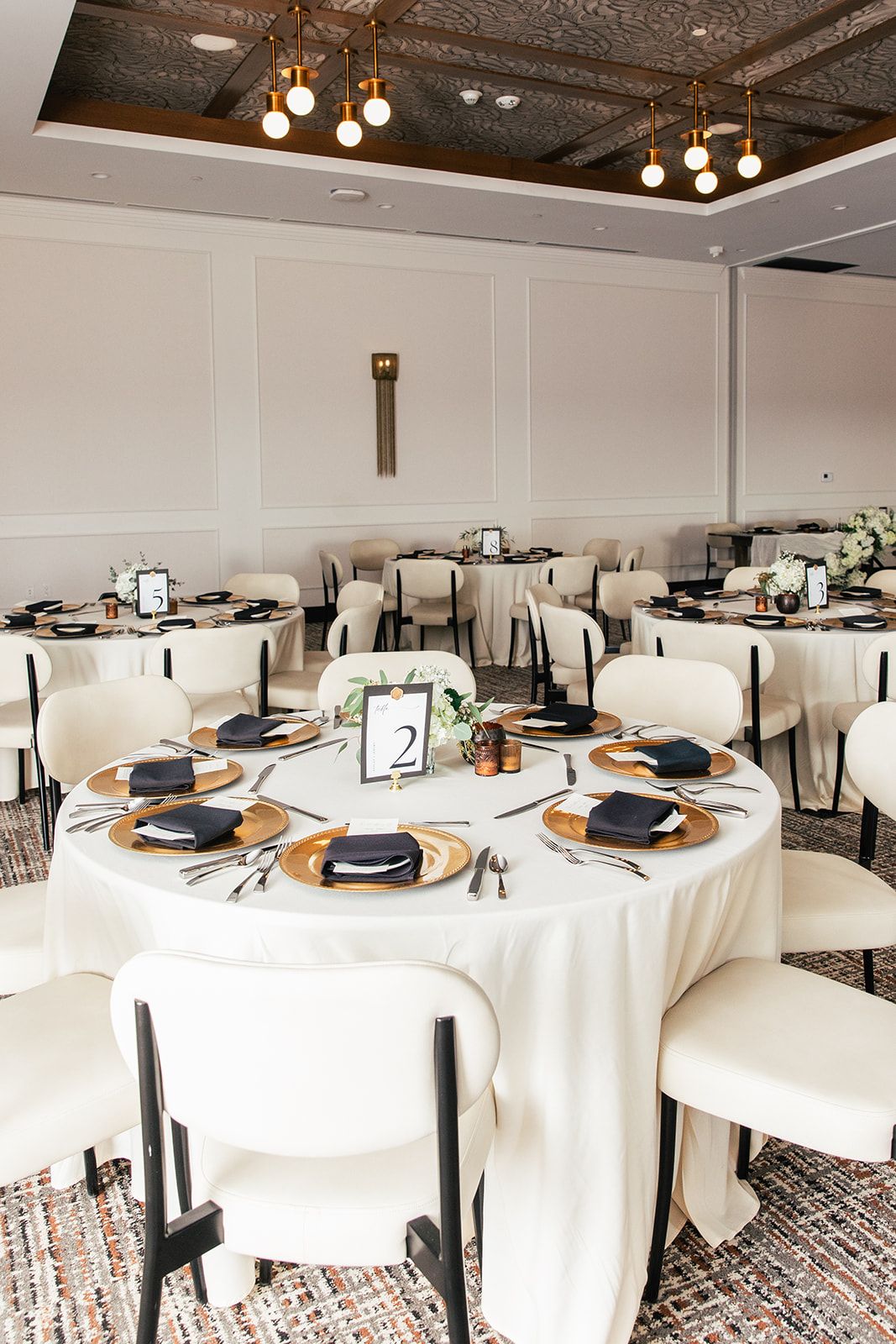 Elegant wedding reception hall with round tables, gold chargers, navy napkins, and warm lighting.