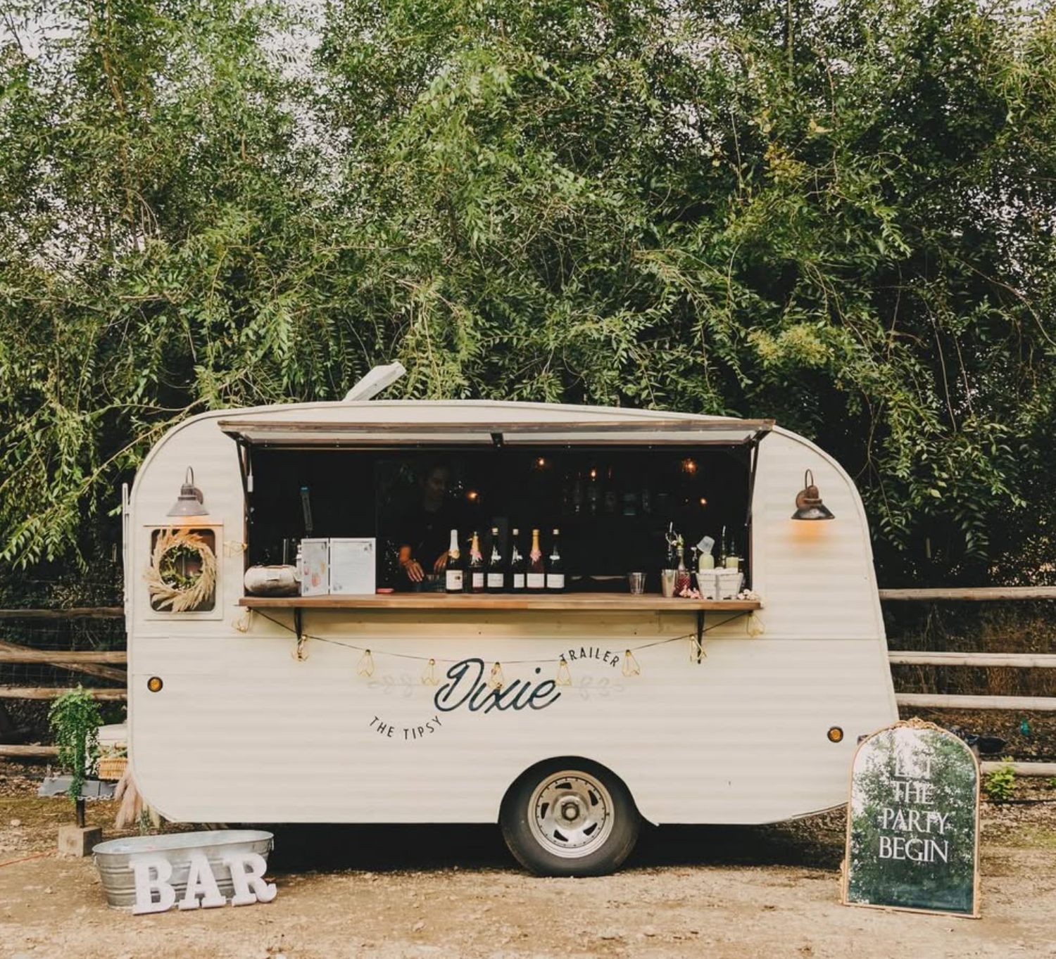 Vintage cream Dixie caravan converted into outdoor mobile bar setup with rustic wooden shelving.