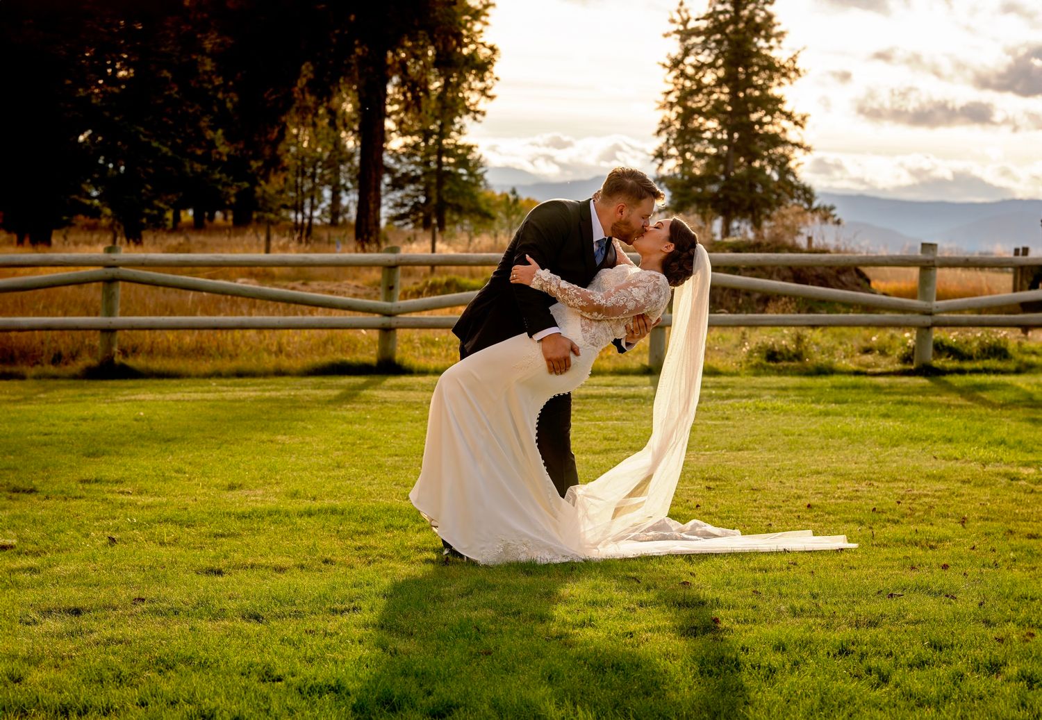 Groom dips bride in elegant white gown on lush green lawn during golden hour with scenic mountains.