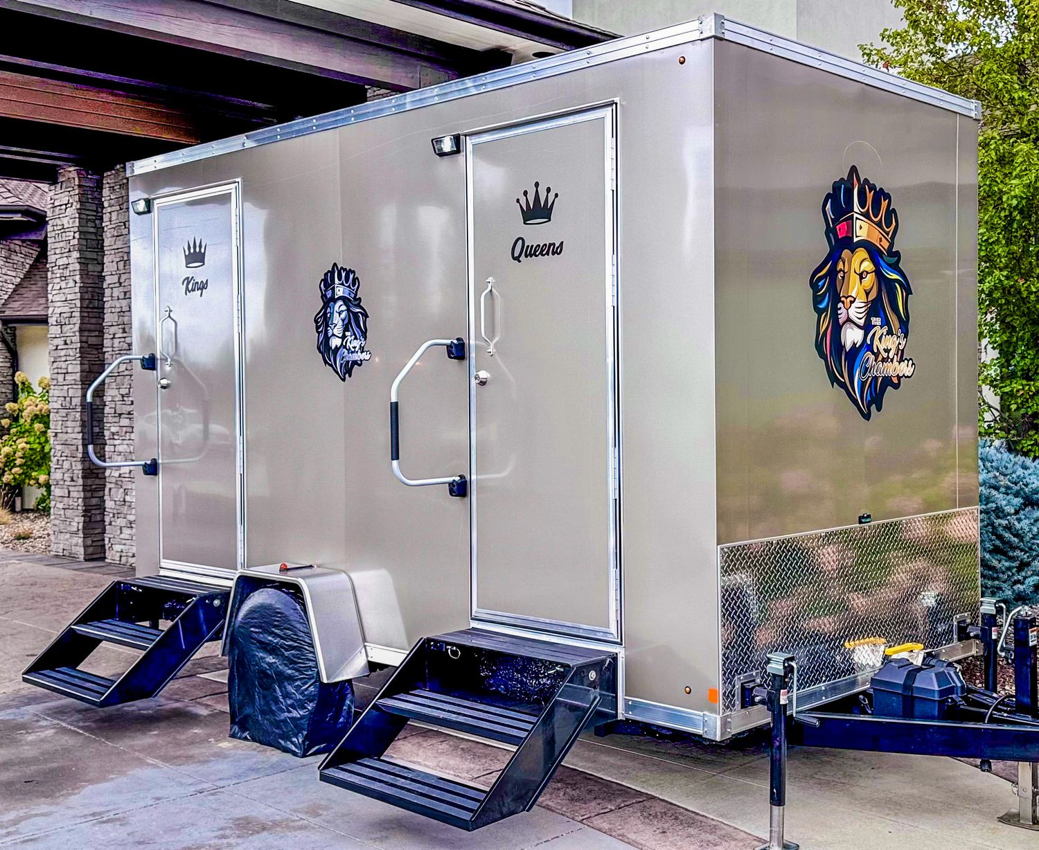 Luxury portable restroom trailer with King and Queen branding, featuring silver exterior and steps, parked outdoors.
