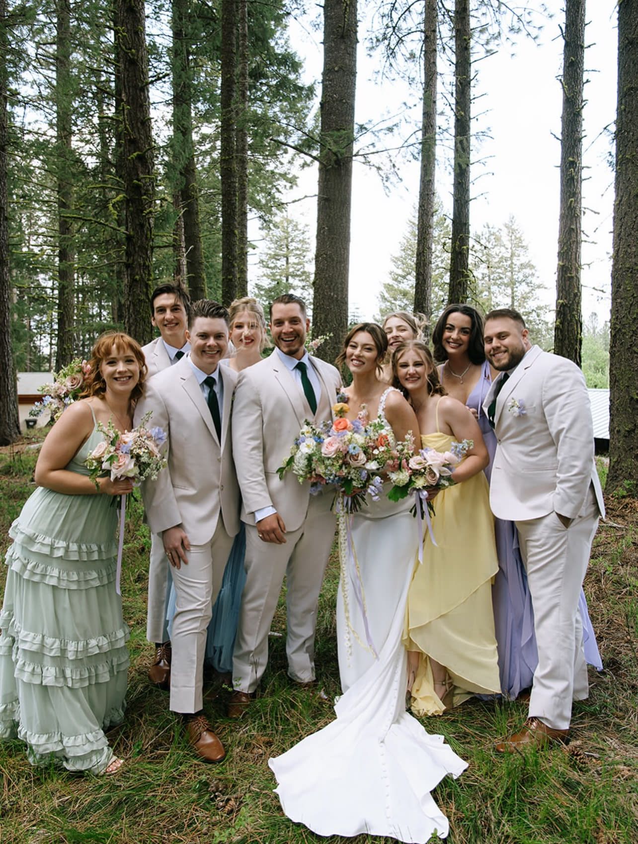 Wedding party posing in forest wearing pastel attire with colorful wildflower bouquets.