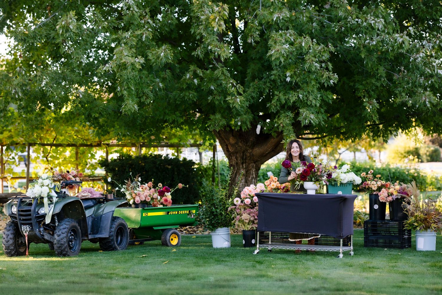 Woman arranging flowers outdoors beside a John Deere utility vehicle under a tree.