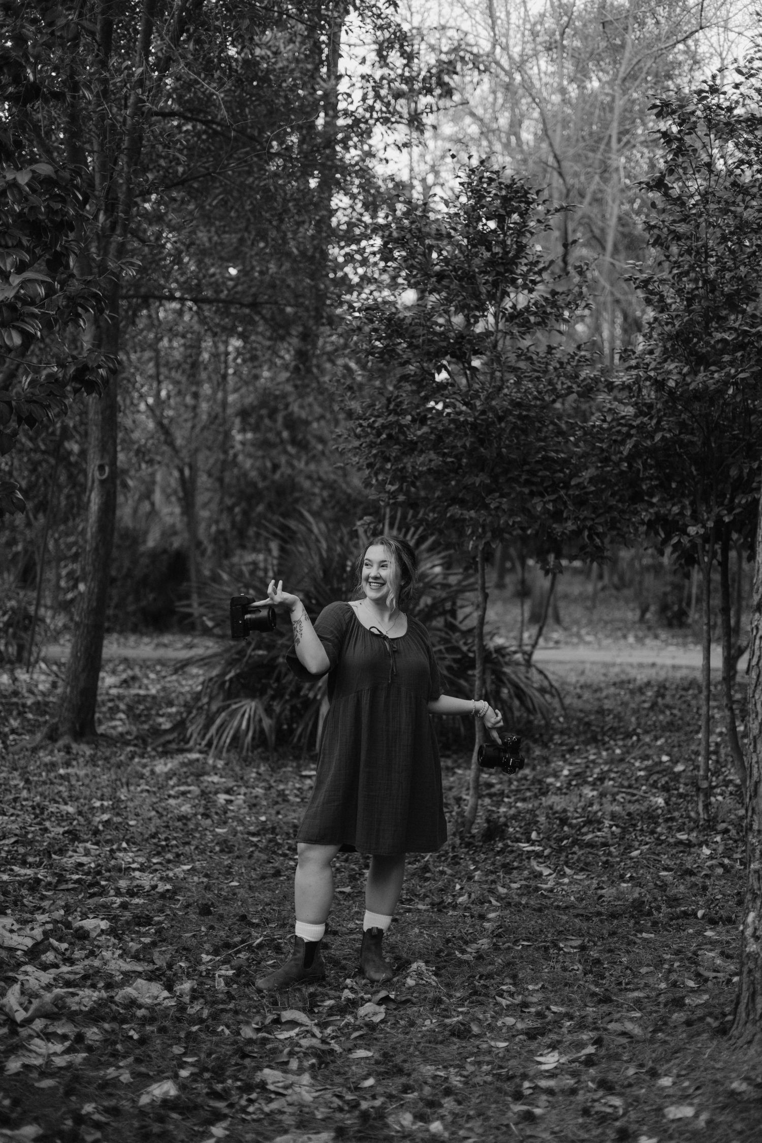 Black and white photo of a smiling woman in a dress holding items while standing on a leaf-covered path in a forest.