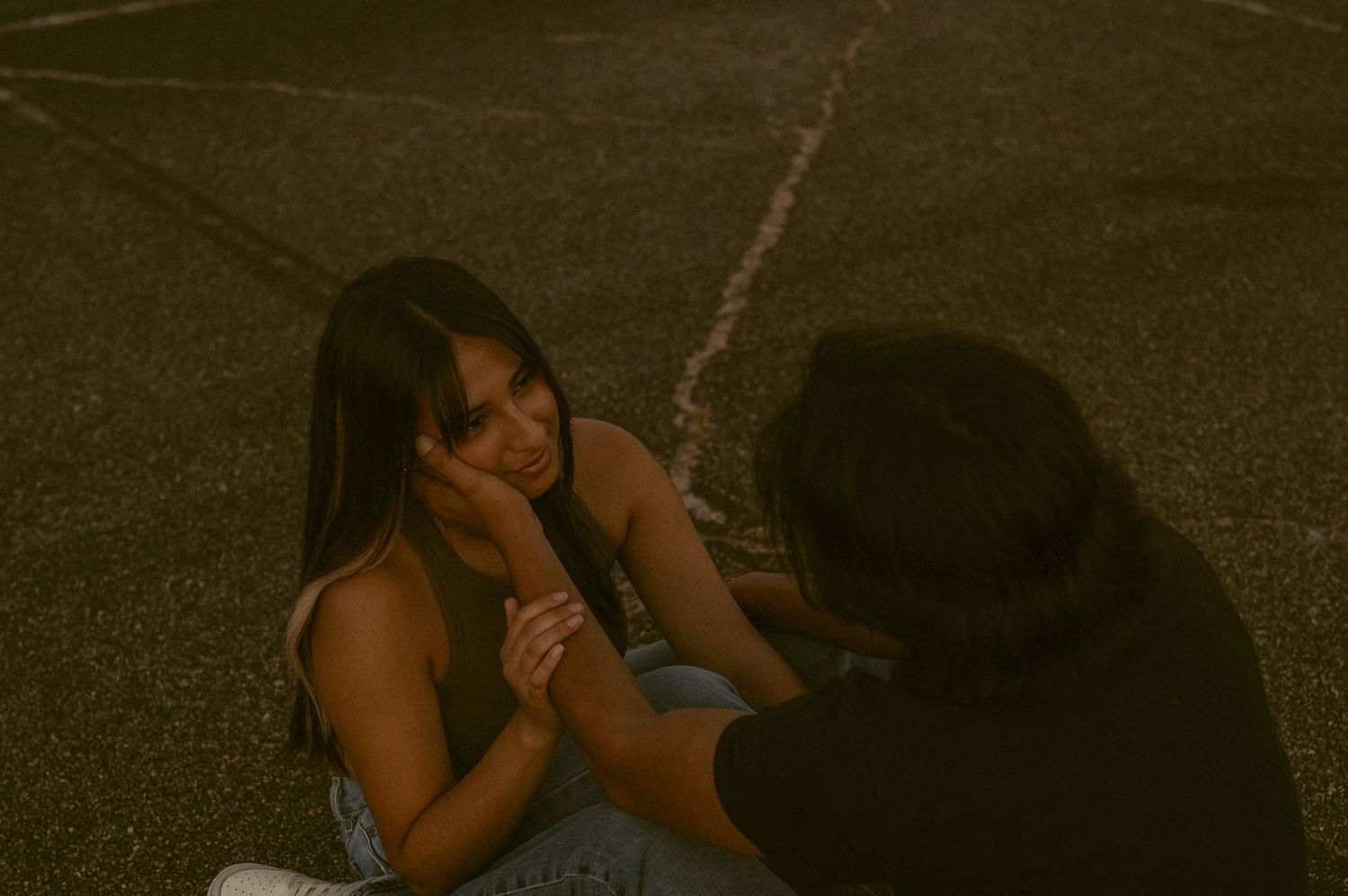 Two people sitting on pavement at dusk, woman resting her cheek on her hand while a person holds her other hand.