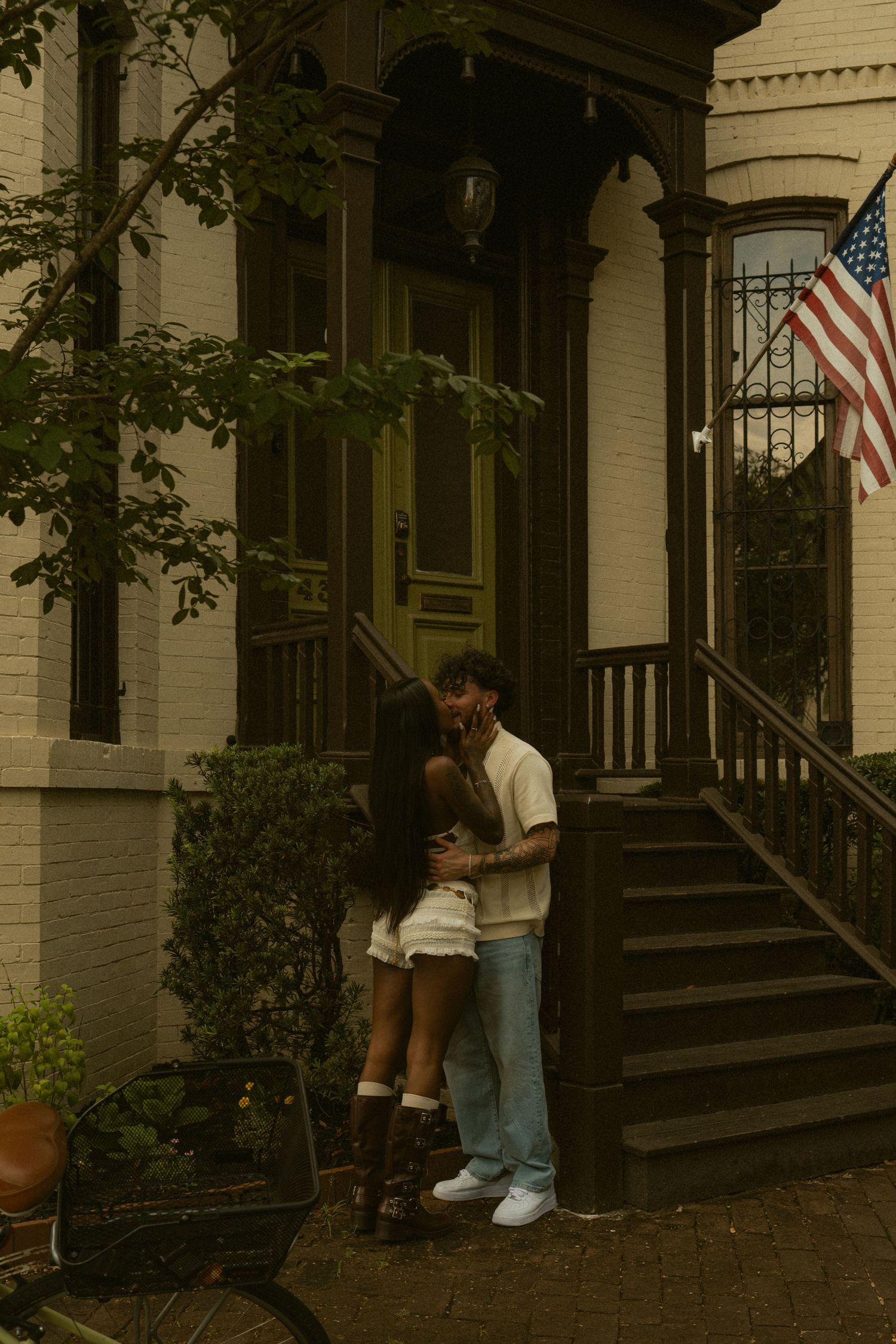 Couple embracing and kissing in front of a brick townhouse with an American flag, surrounded by lush greenery.