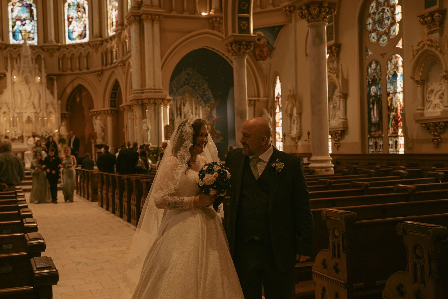 Bride in white gown and veil walks down church aisle with older man in suit during wedding ceremony.