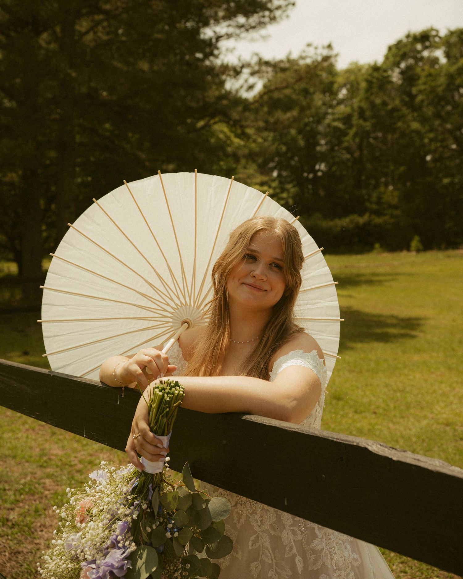 Smiling bride in white lace dress holds floral bouquet and parasol umbrella outdoors in lush green park setting.