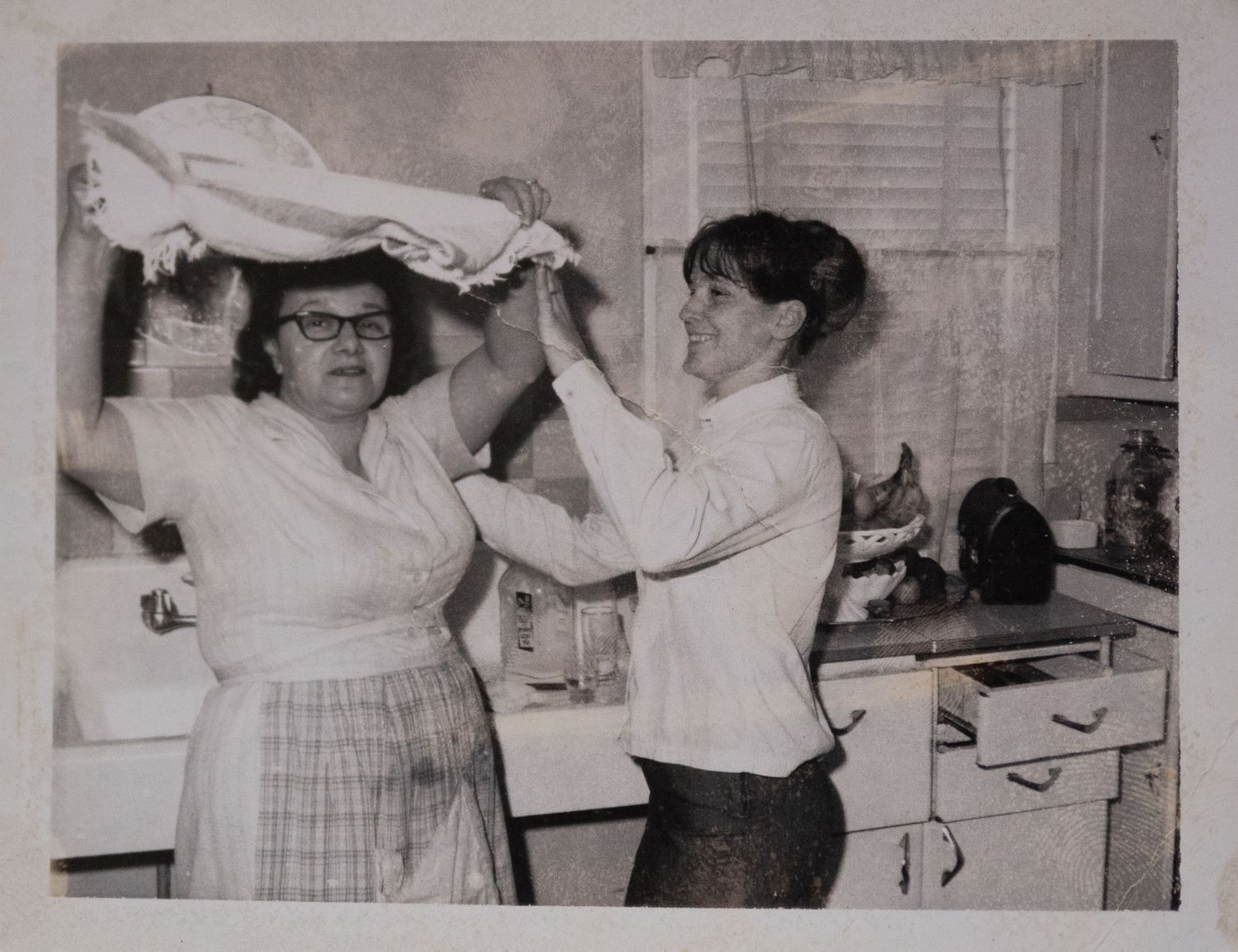 Two women folding laundry together in a vintage kitchen, black and white photo from mid-20th century.