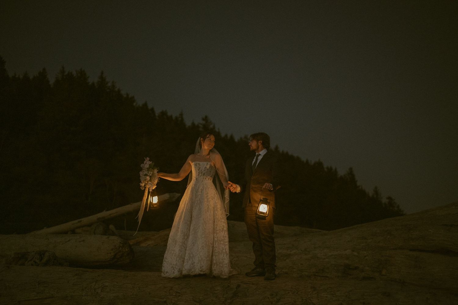 Bride and groom holding lanterns at night with dark forest backdrop during romantic outdoor wedding ceremony.