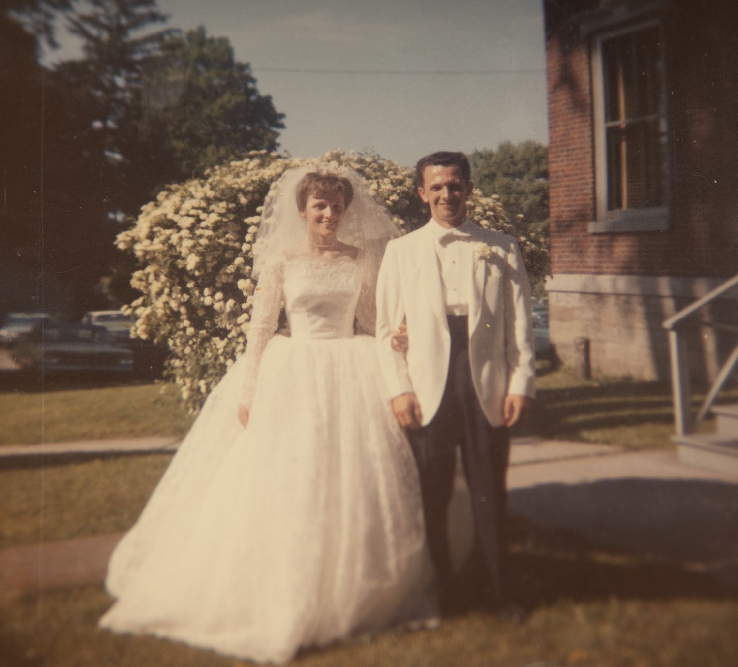Vintage 1950s wedding photo of bride in white gown and groom in white tuxedo posing outdoors near flowering bushes.