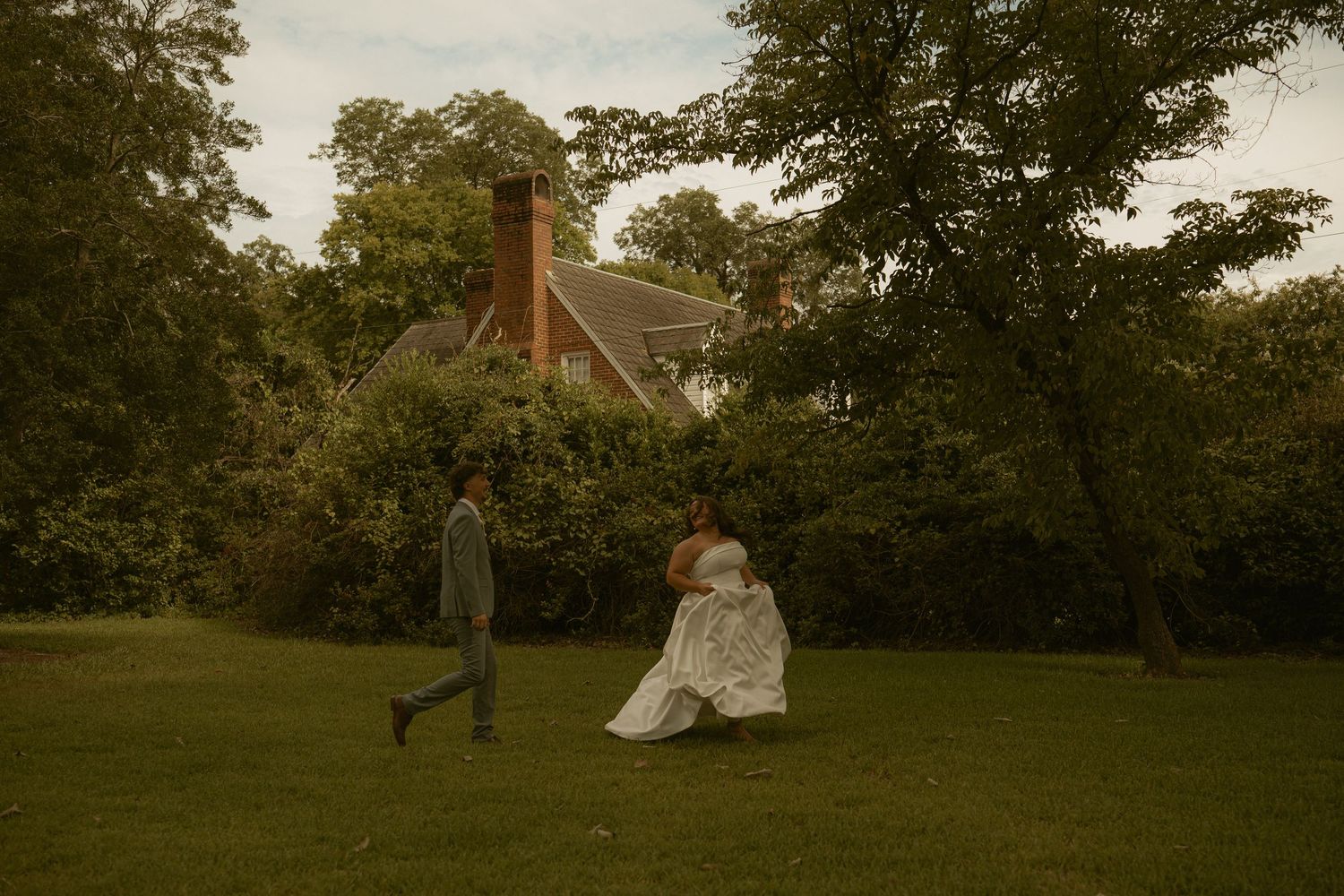 Bride in white gown and groom in suit walk across lush green lawn near historic brick manor surrounded by trees.