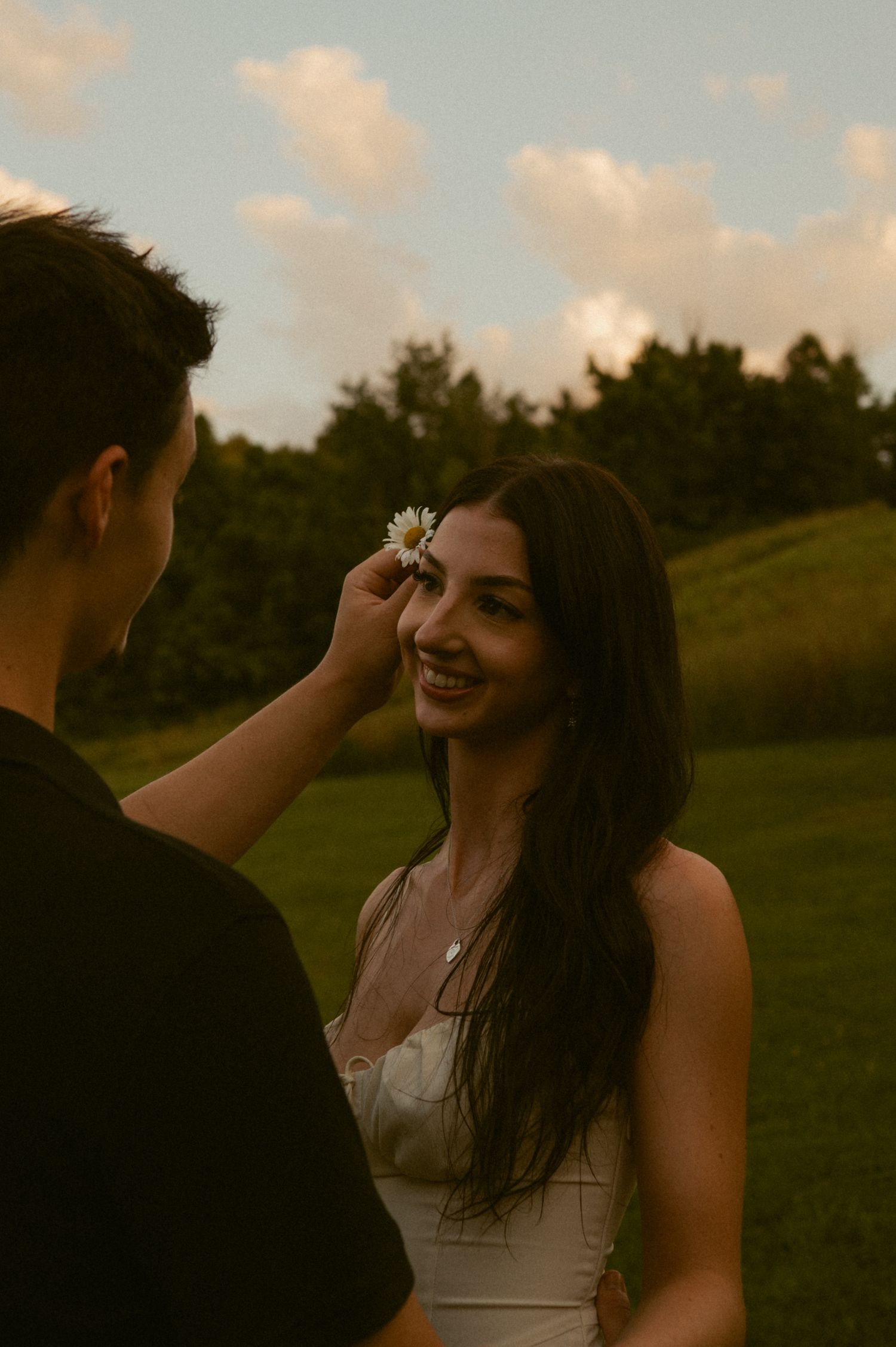 A man places a daisy flower in a smiling woman's hair outdoors at golden hour.