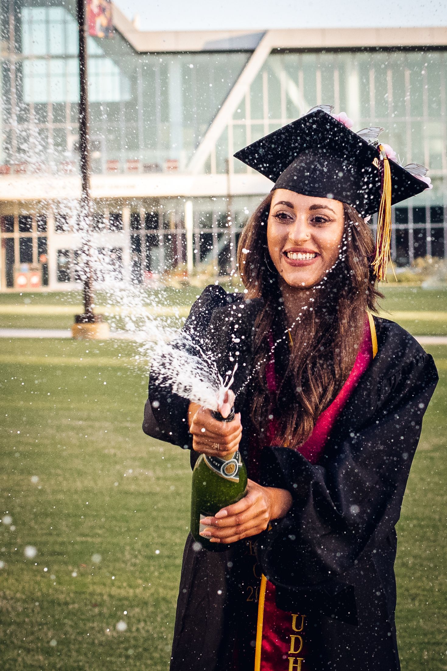 Graduate celebrates with champagne spray on campus lawn.