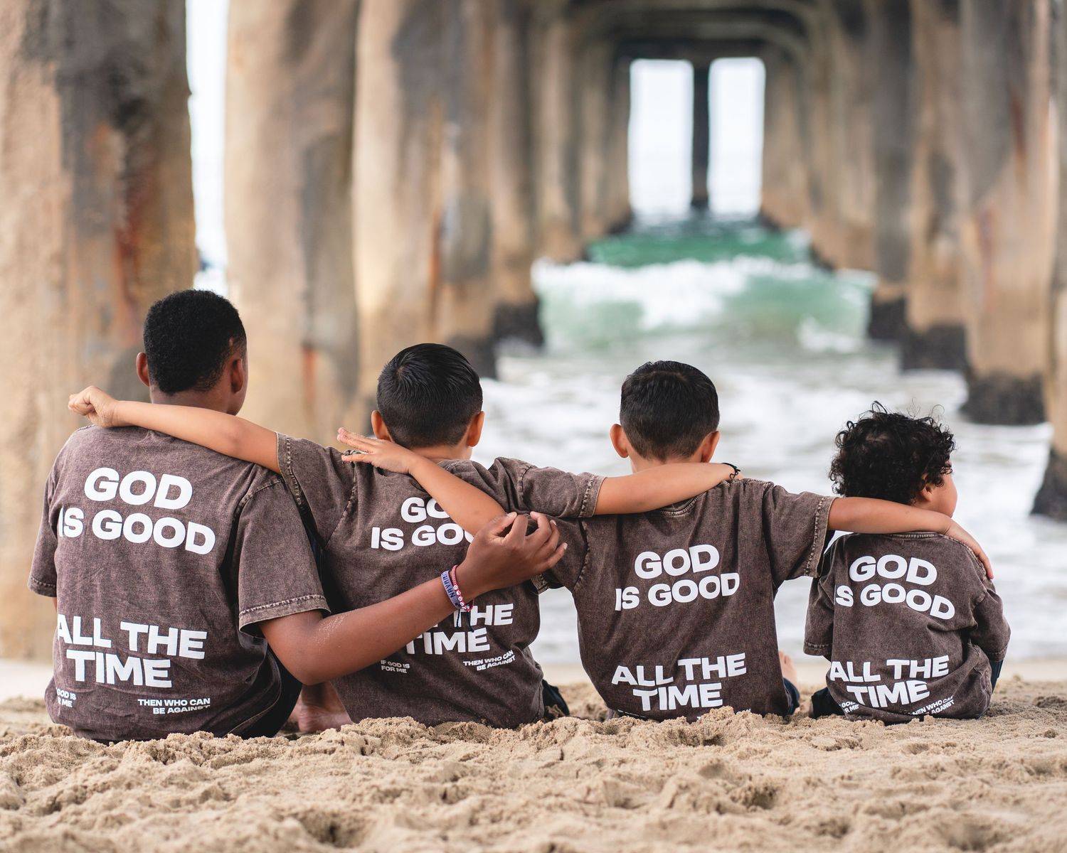 Four children sit on beach sand under a pier, wearing matching brown 'God Is Good All The Time' t-shirts.