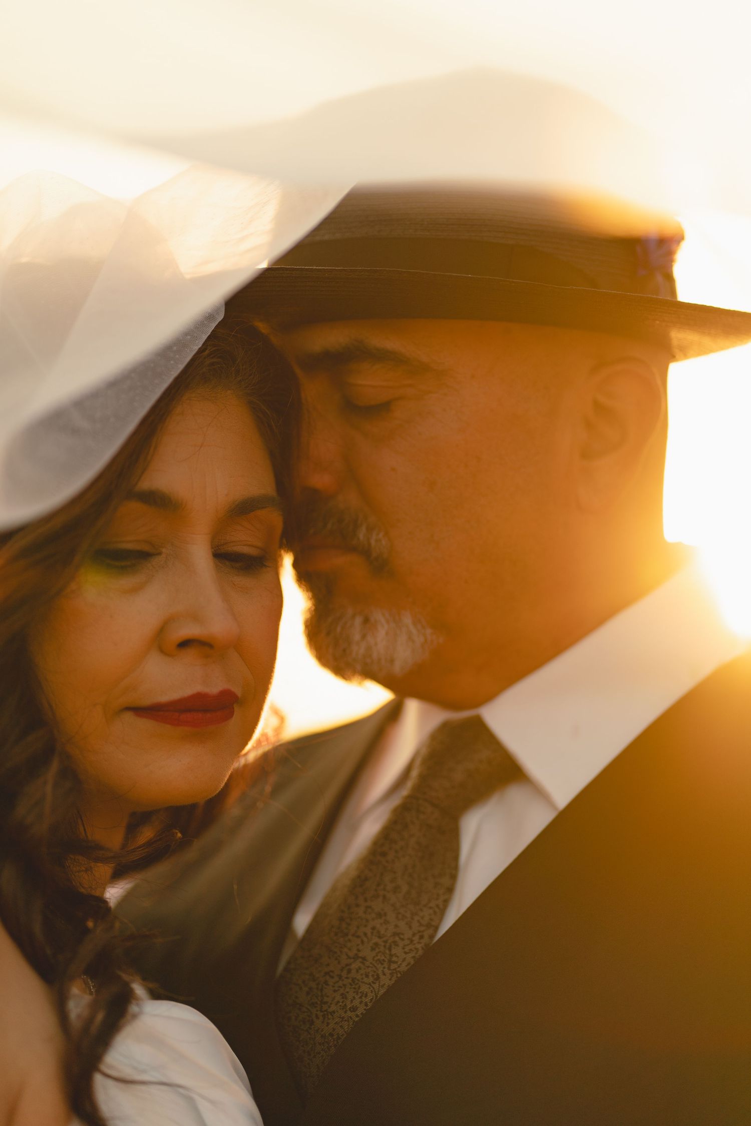 A couple shares a romantic moment at golden hour, the man in a fedora nuzzling close to a woman in a white veil.