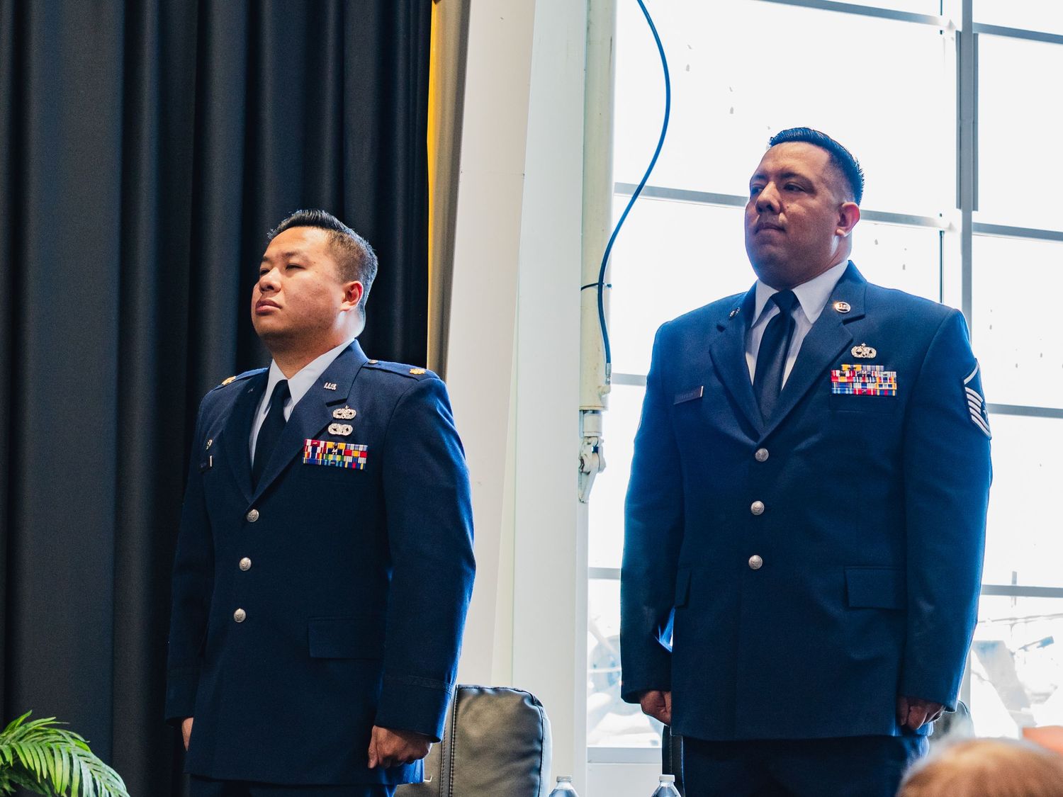 Two US Air Force military personnel in dress blue uniforms standing at attention during a formal ceremony indoors.