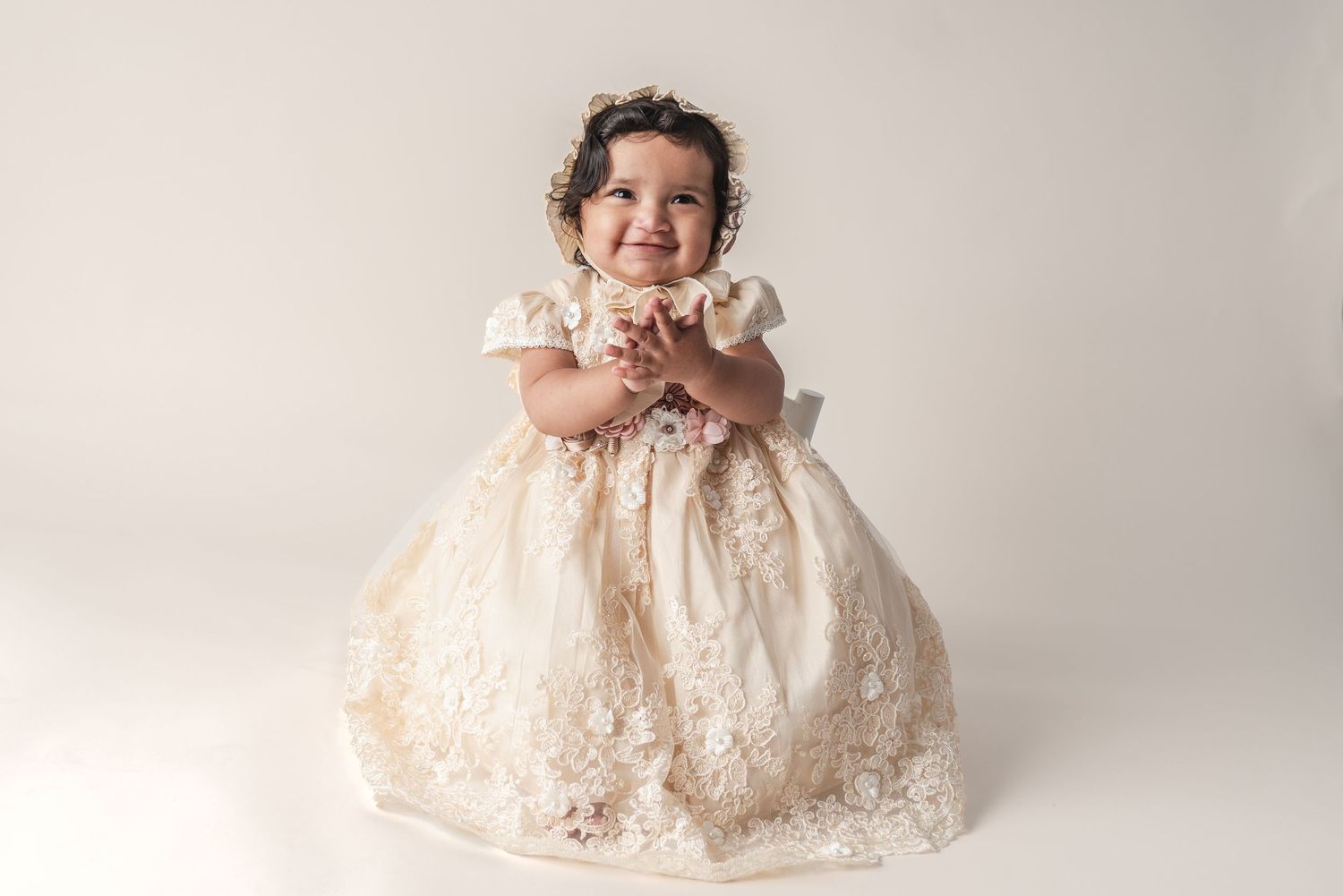Baby wearing vintage lace dress poses on cream backdrop for professional portrait.
