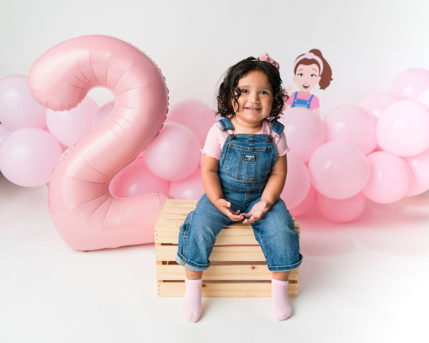 Birthday setup with pink number 2 balloon and child sitting on wooden crate.