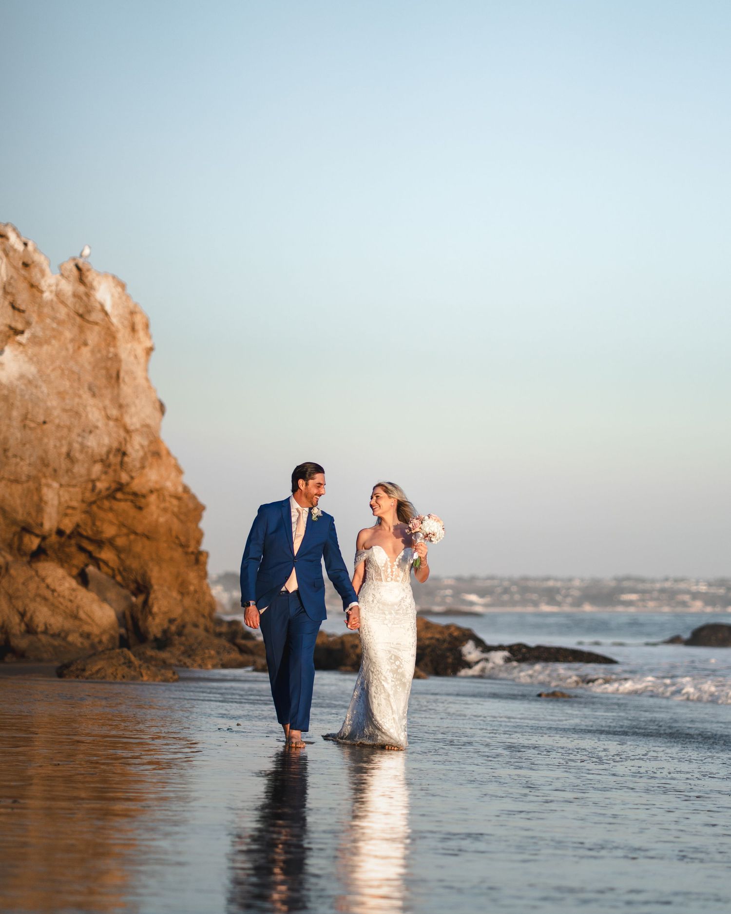 A couple in wedding attire poses for romantic sunset photos along a rocky beach shoreline.