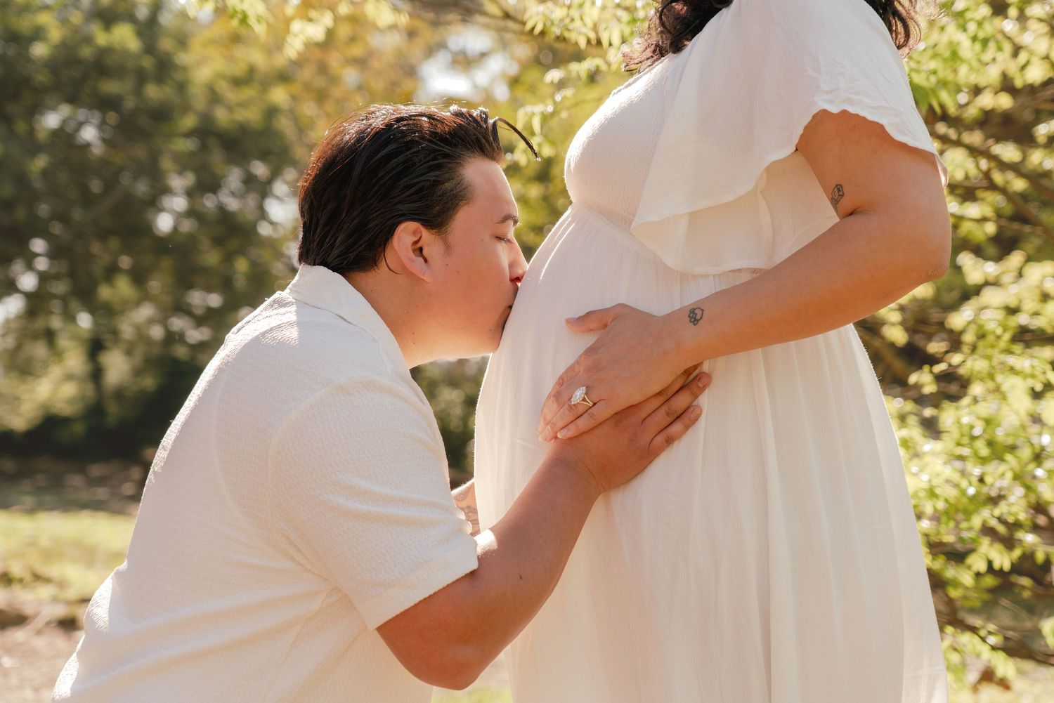 A couple in white wedding attire embraces in a sun-dappled outdoor setting during a romantic photoshoot.