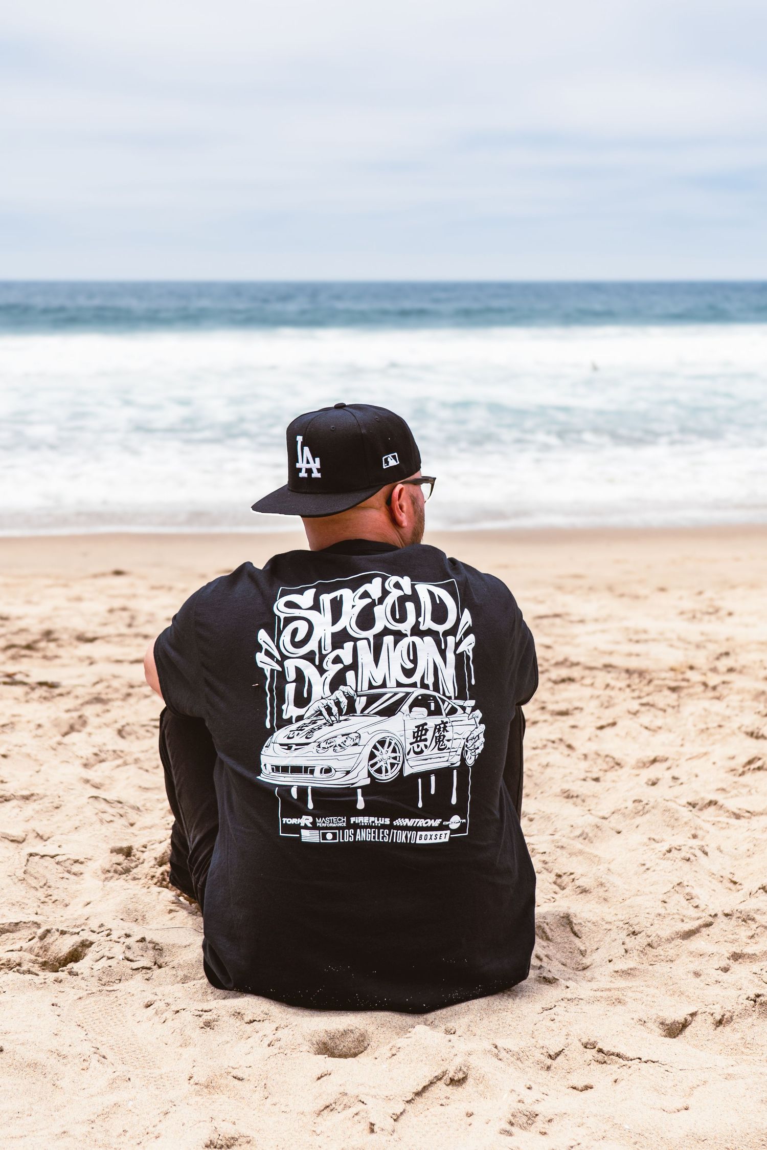 Person wearing LA Dodgers cap and black Speed Demon graphic sweatshirt sits on sandy beach overlooking ocean waves.