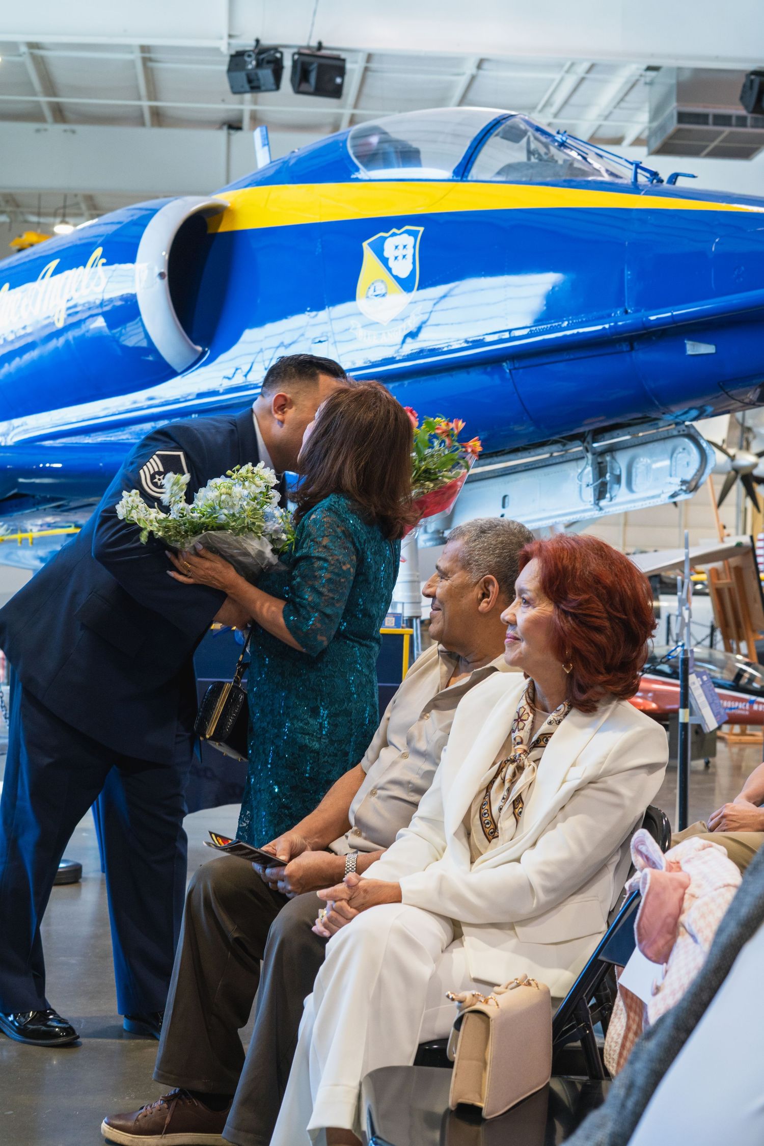 Man presenting flowers to woman at Blue Angels jet display, with seated guests watching at aviation museum ceremony.