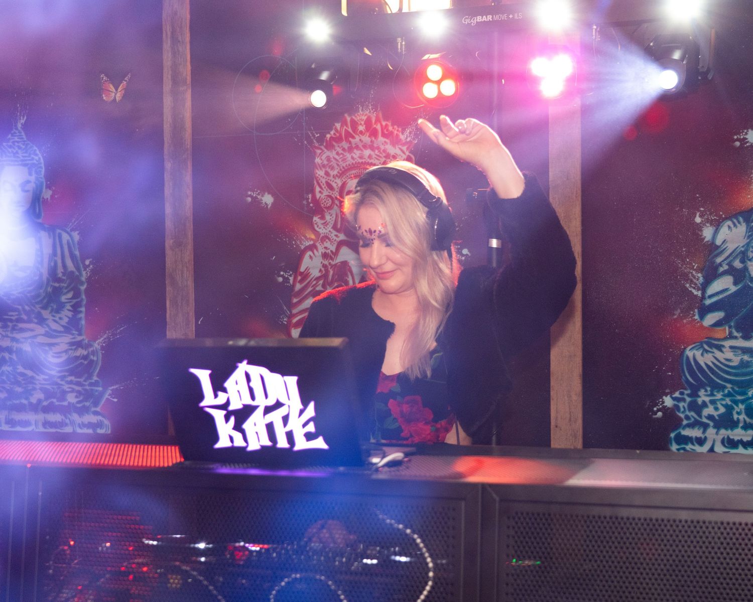 DJ Lady Kate performing at a nightclub with colorful stage lighting and artistic backdrop decorations.