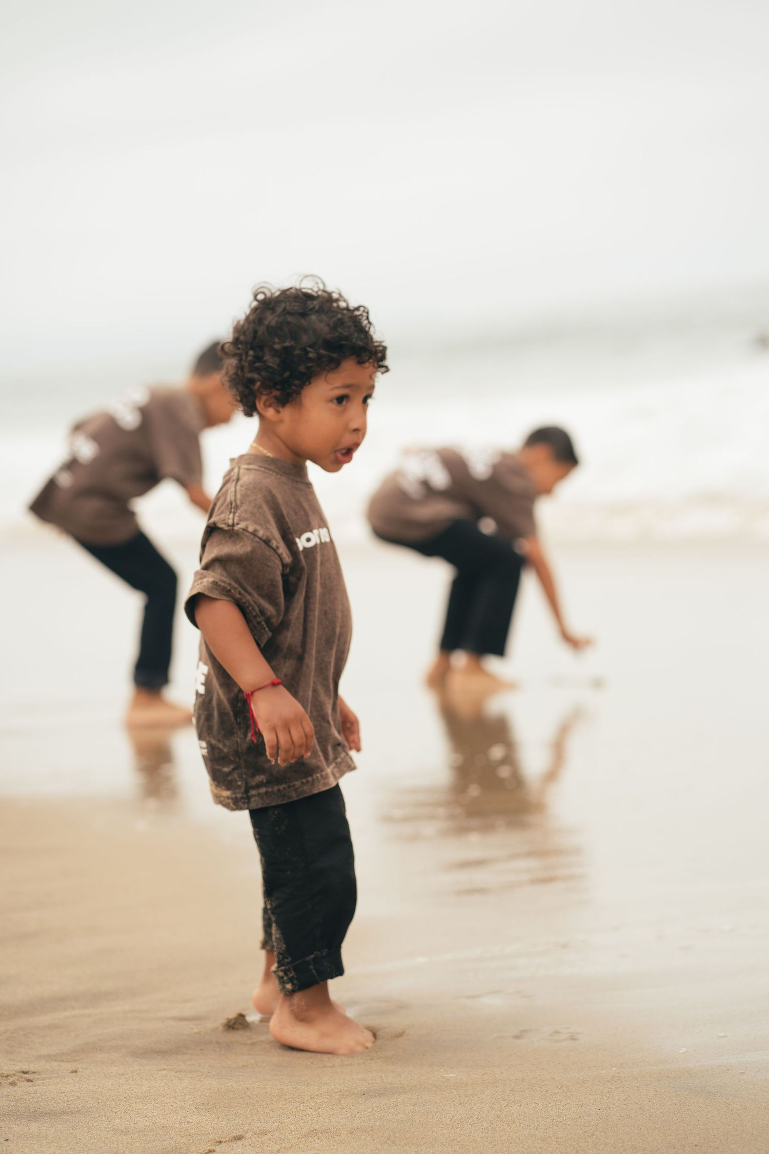Young curly-haired child stands barefoot on a sandy beach while two others play in the shallow water behind him.