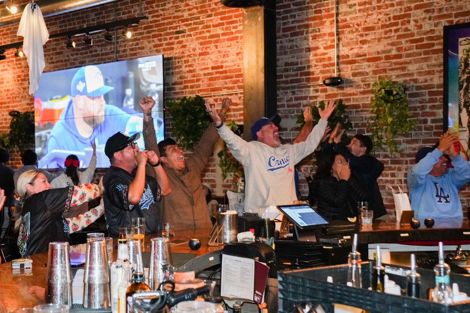 Excited sports fans cheer at a bar with brick walls, TV screens, and drinks during a lively game-watching event.