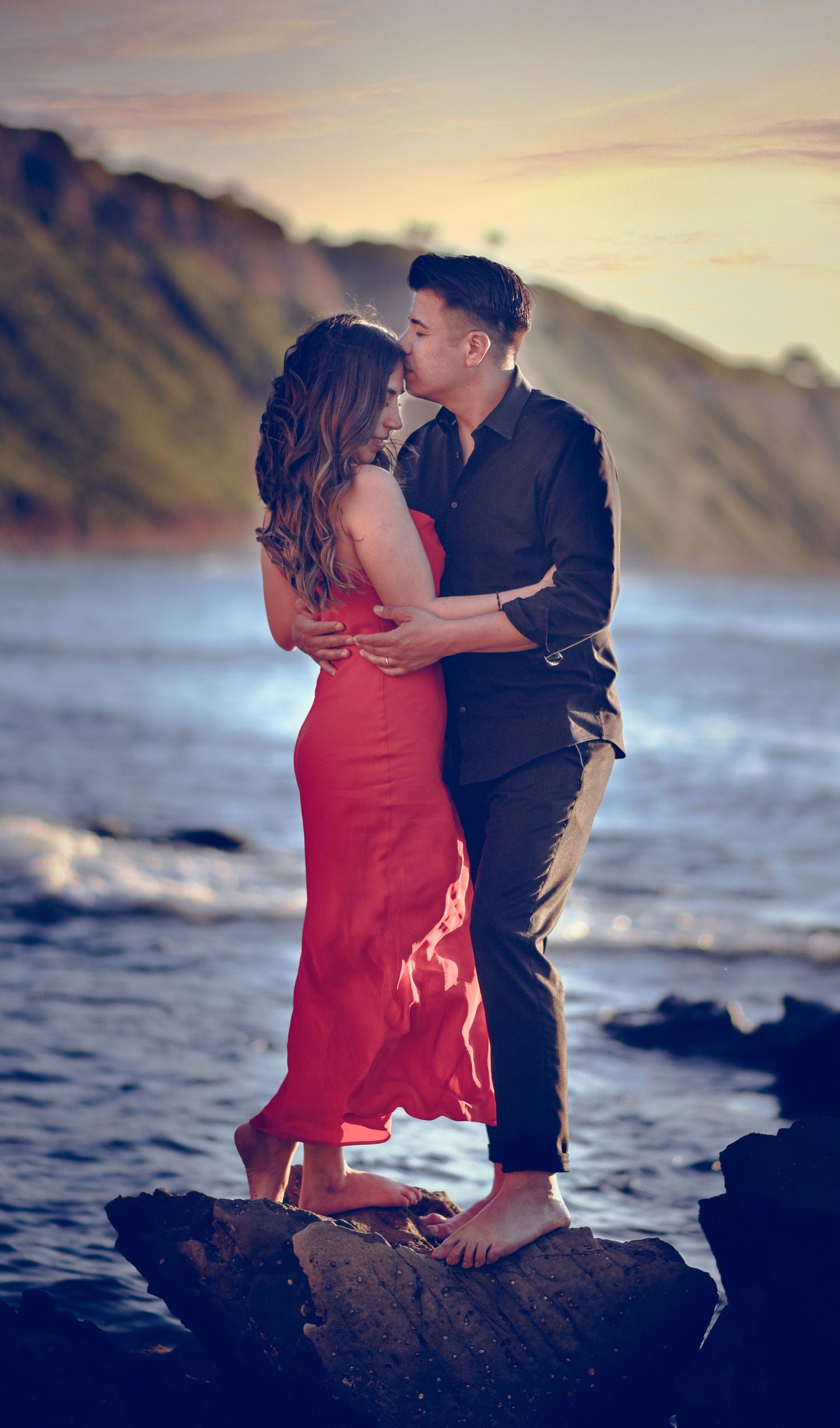 A romantic couple shares an intimate moment on coastal rocks at sunset with mountains and ocean waves in the background.