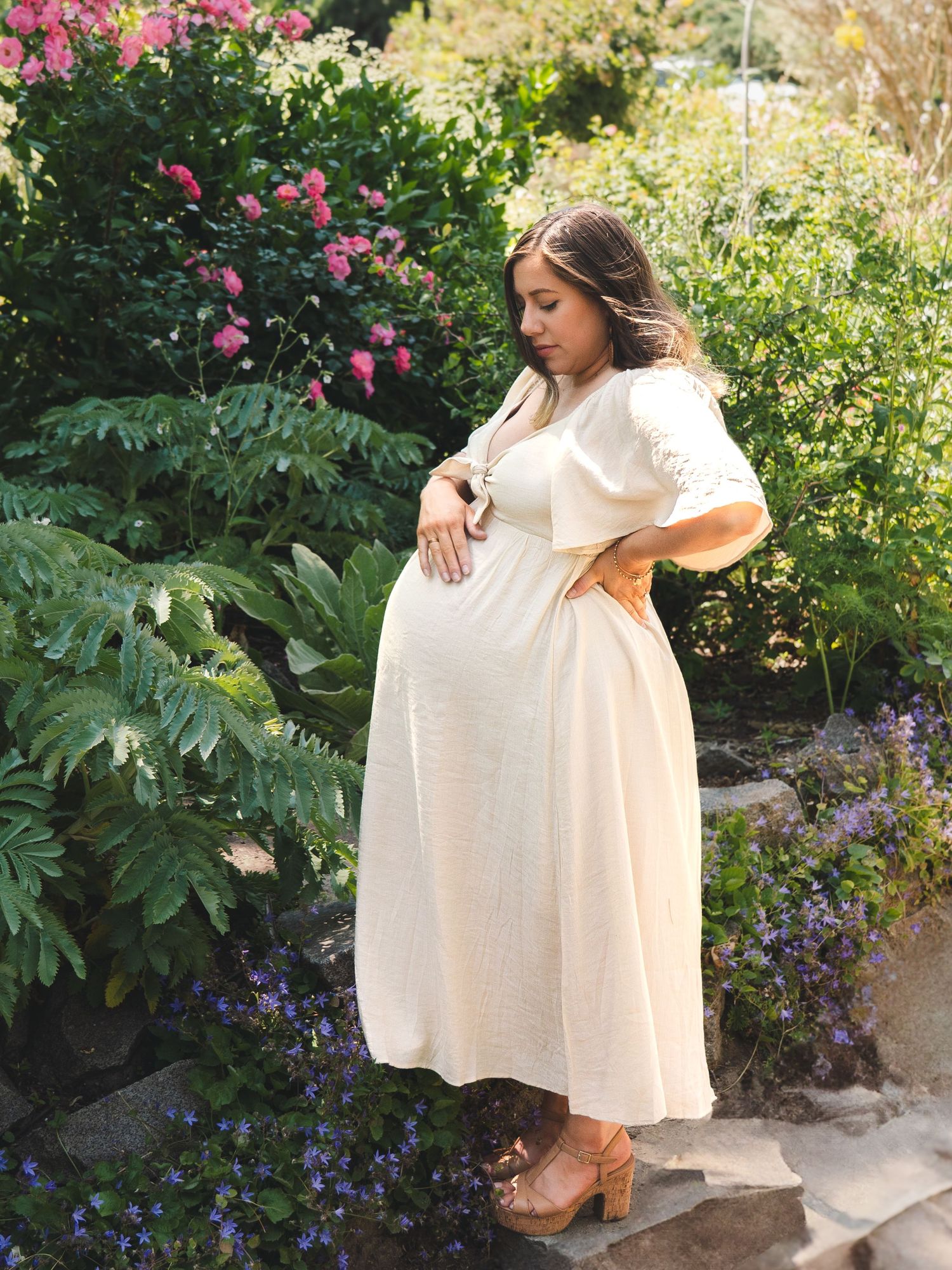 Pregnant woman in white dress poses in garden with pink flowers and purple blooms.