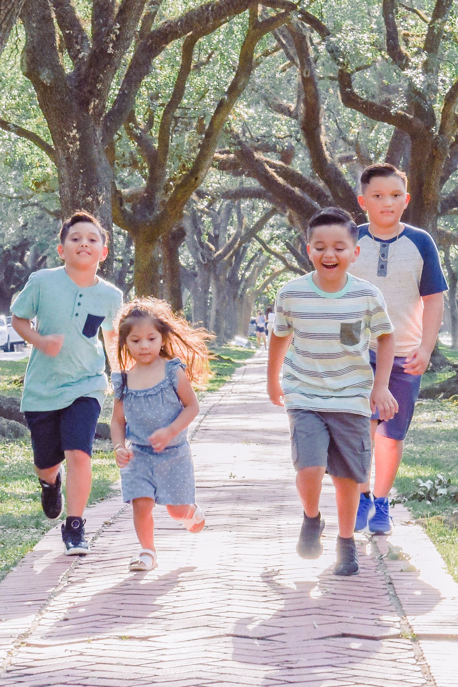 Young children running and playing together on a brick pathway under shady trees.
