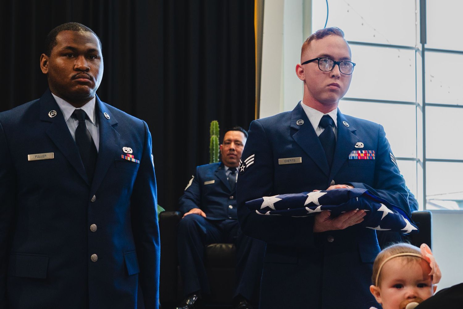 Two Air Force servicemen in dress uniforms hold a folded flag at a military ceremony.