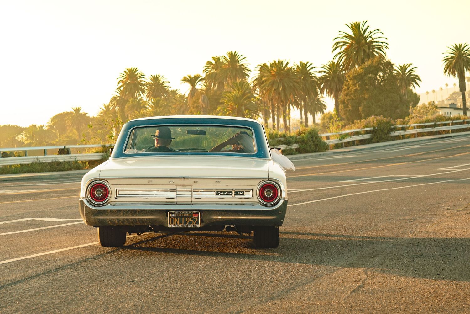 Vintage white Ford Falcon driving on a sunny road lined with palm trees at golden hour.