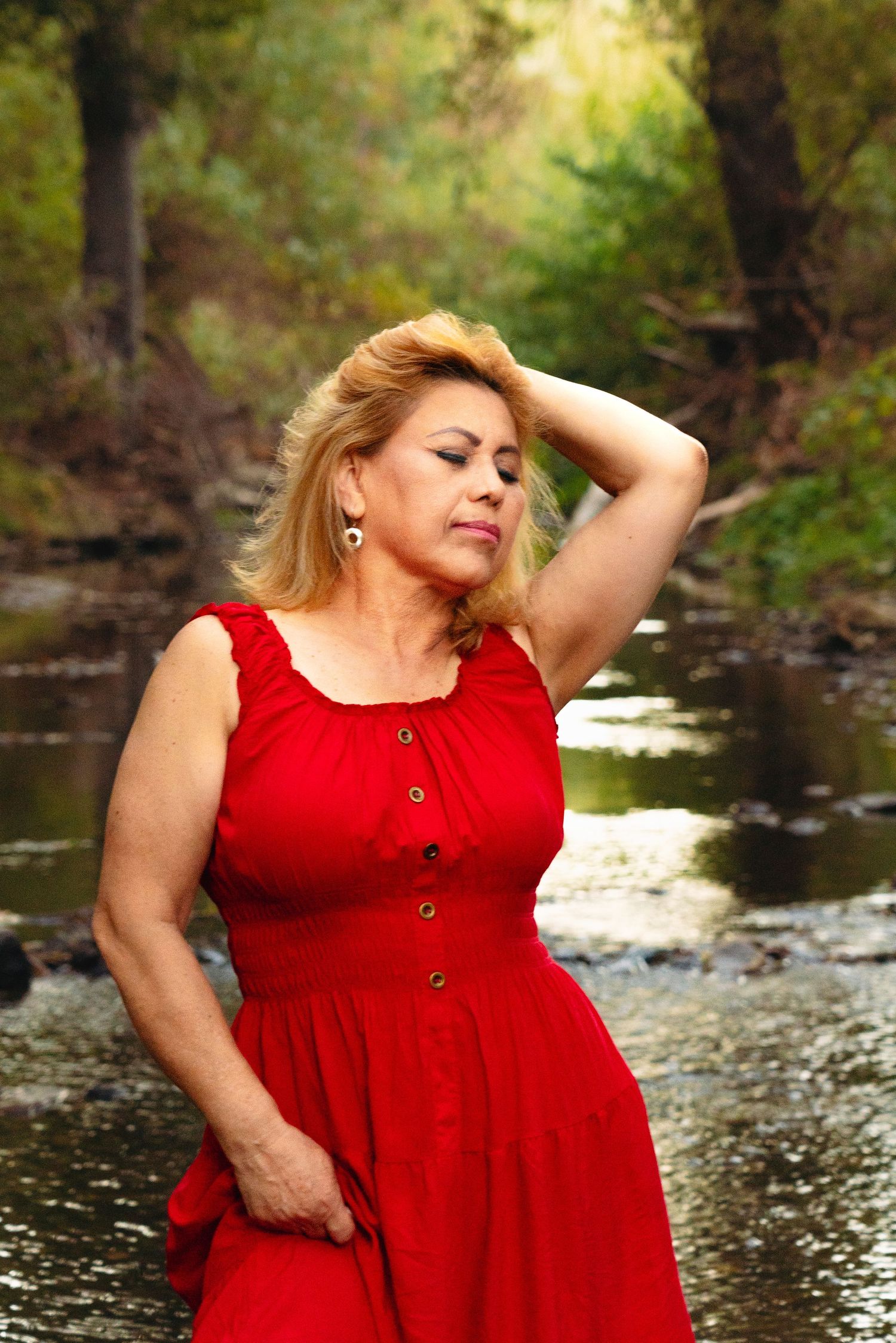A woman in a vibrant red dress poses thoughtfully by a serene creek in a wooded setting.