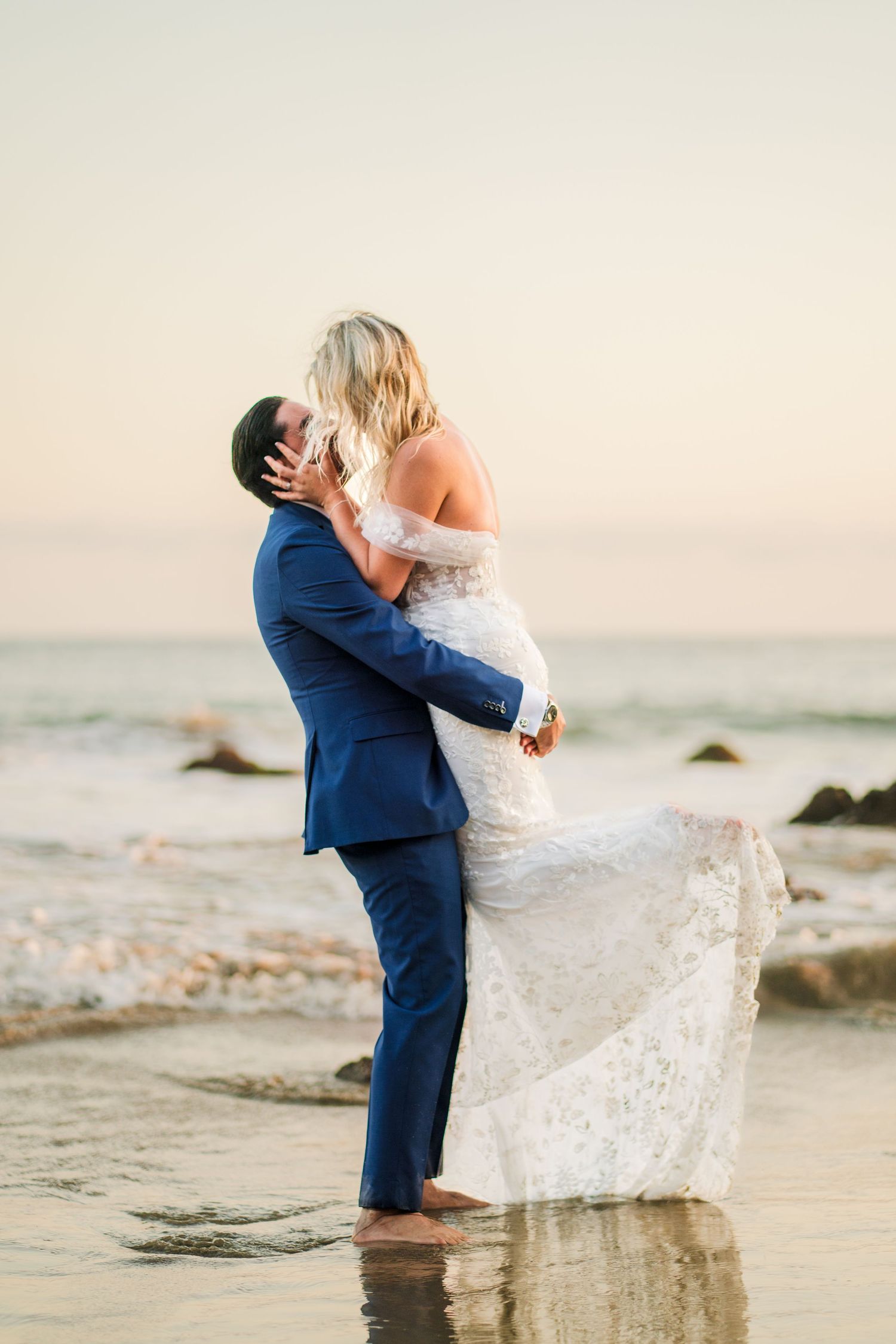 A couple in wedding attire embrace and twirl on a beach at sunset, creating a romantic seaside moment.