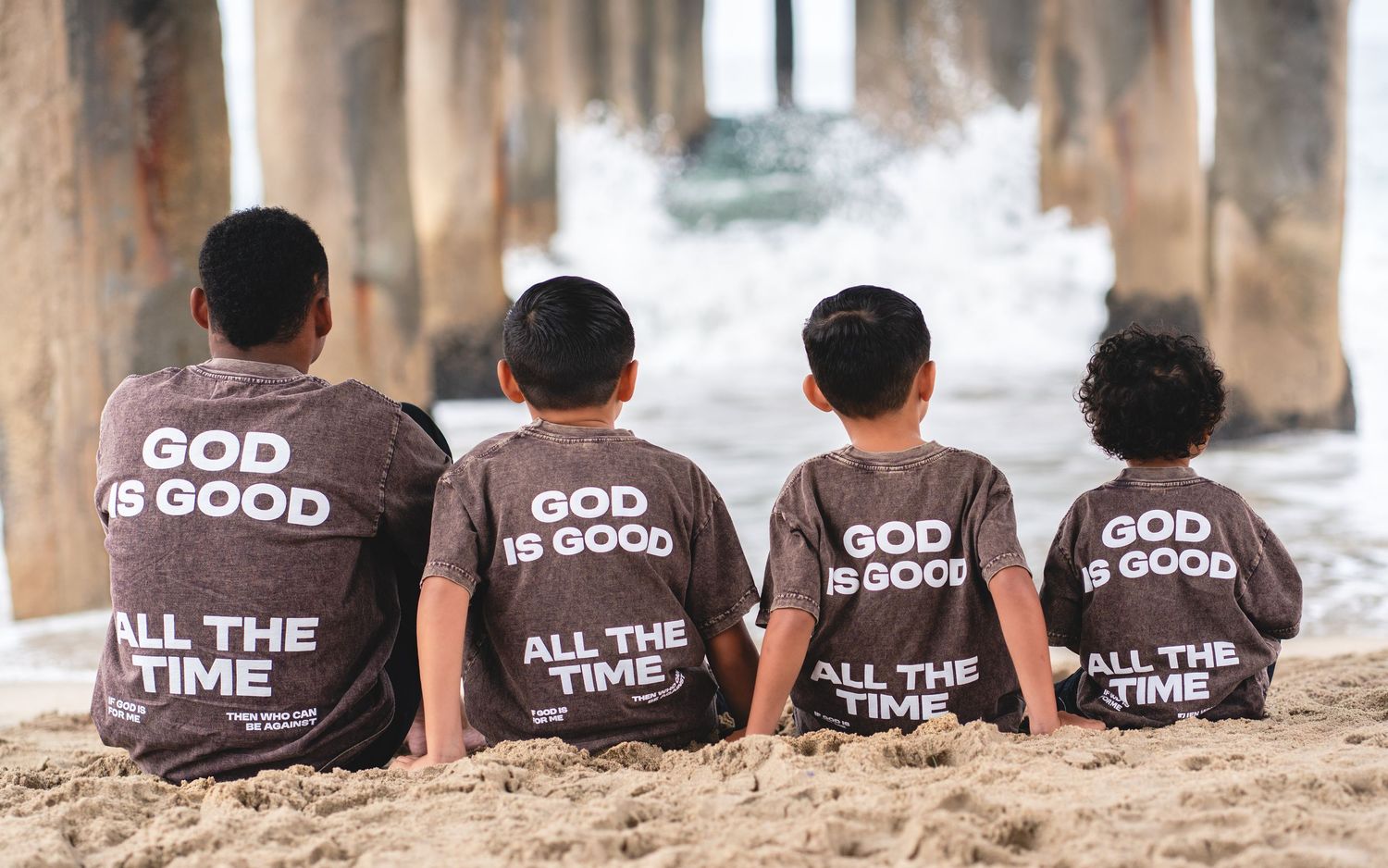 Four children sit on sandy beach wearing matching gray t-shirts reading God Is Good All The Time near ocean pier.
