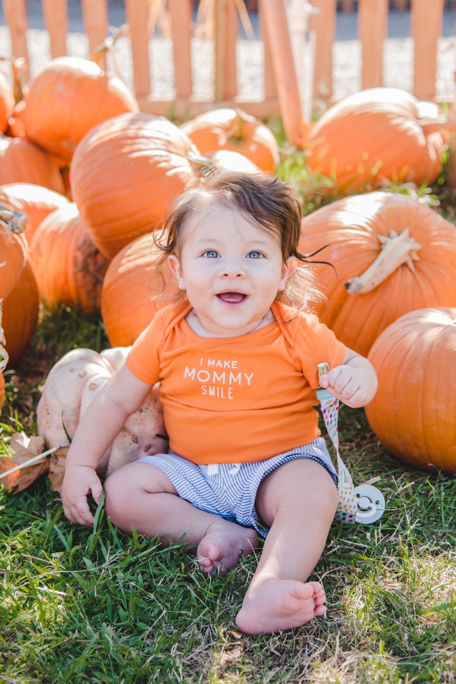 Adorable baby in orange onesie sitting among large pumpkins on grassy ground.