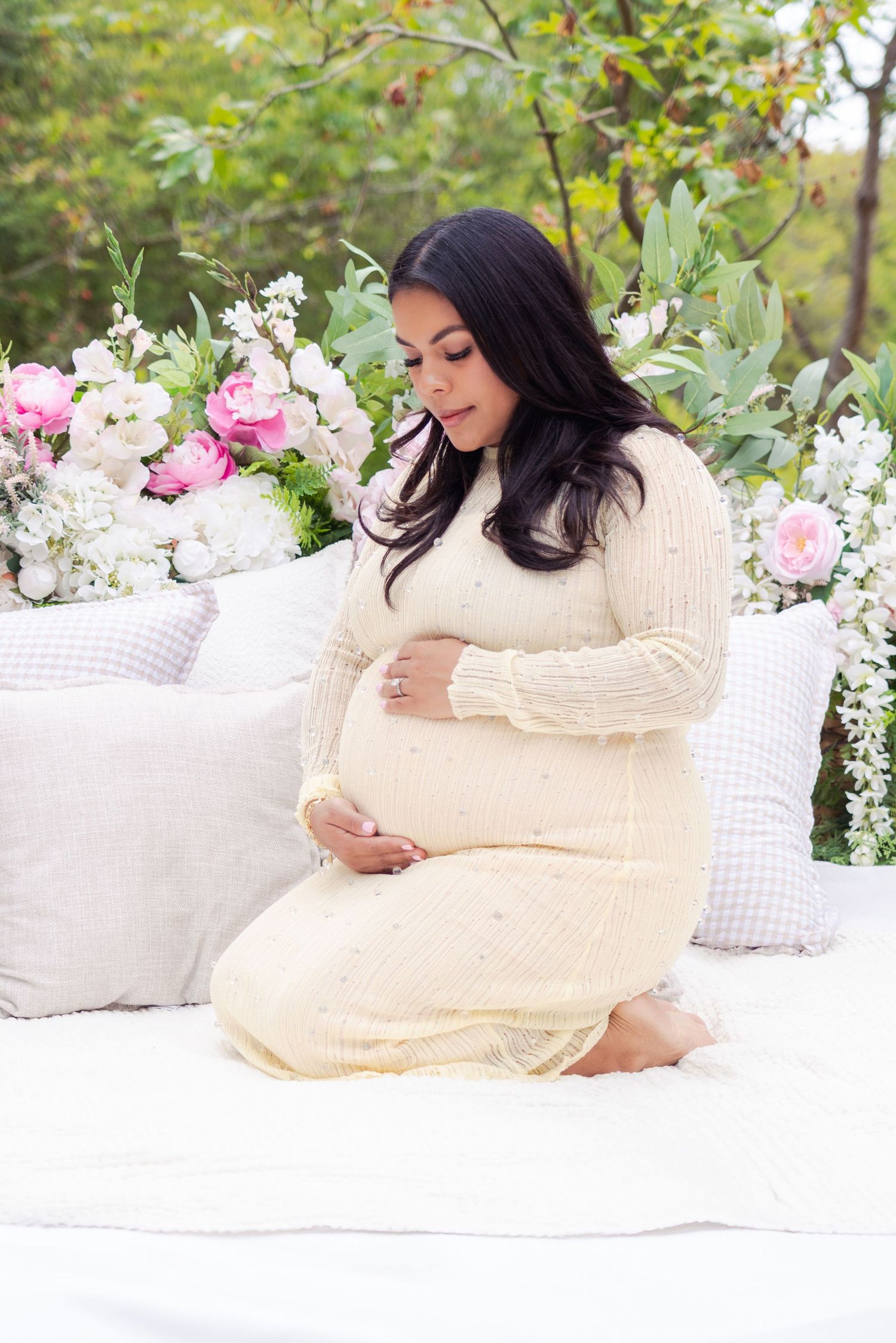 Maternity photos on white bedding with pink and white flowers in garden setting.