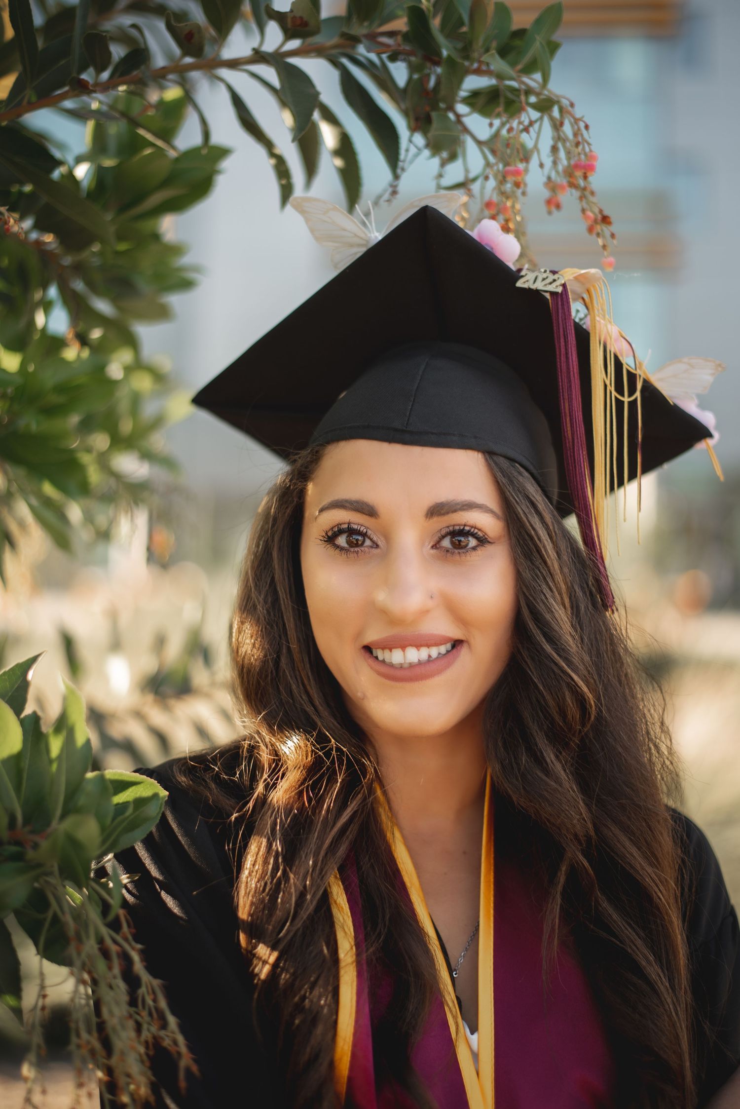 Graduate in cap and burgundy gown poses outdoors among greenery.