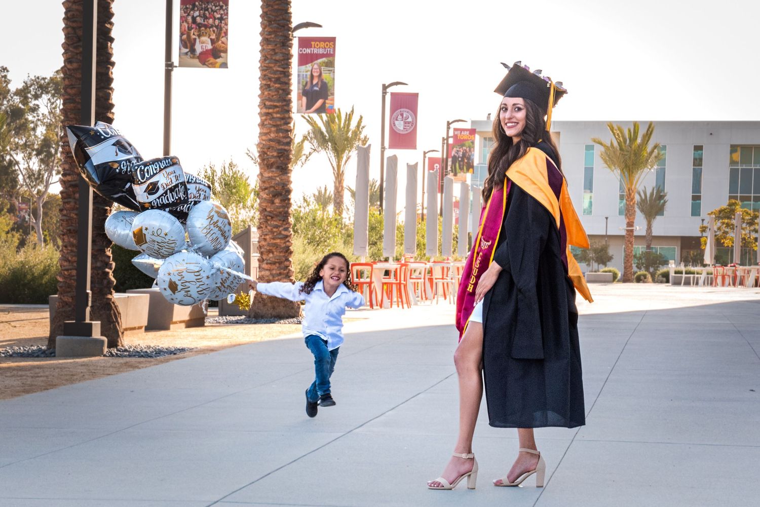 Graduate in cap and gown poses with a small child running in front of palm trees on a university campus.